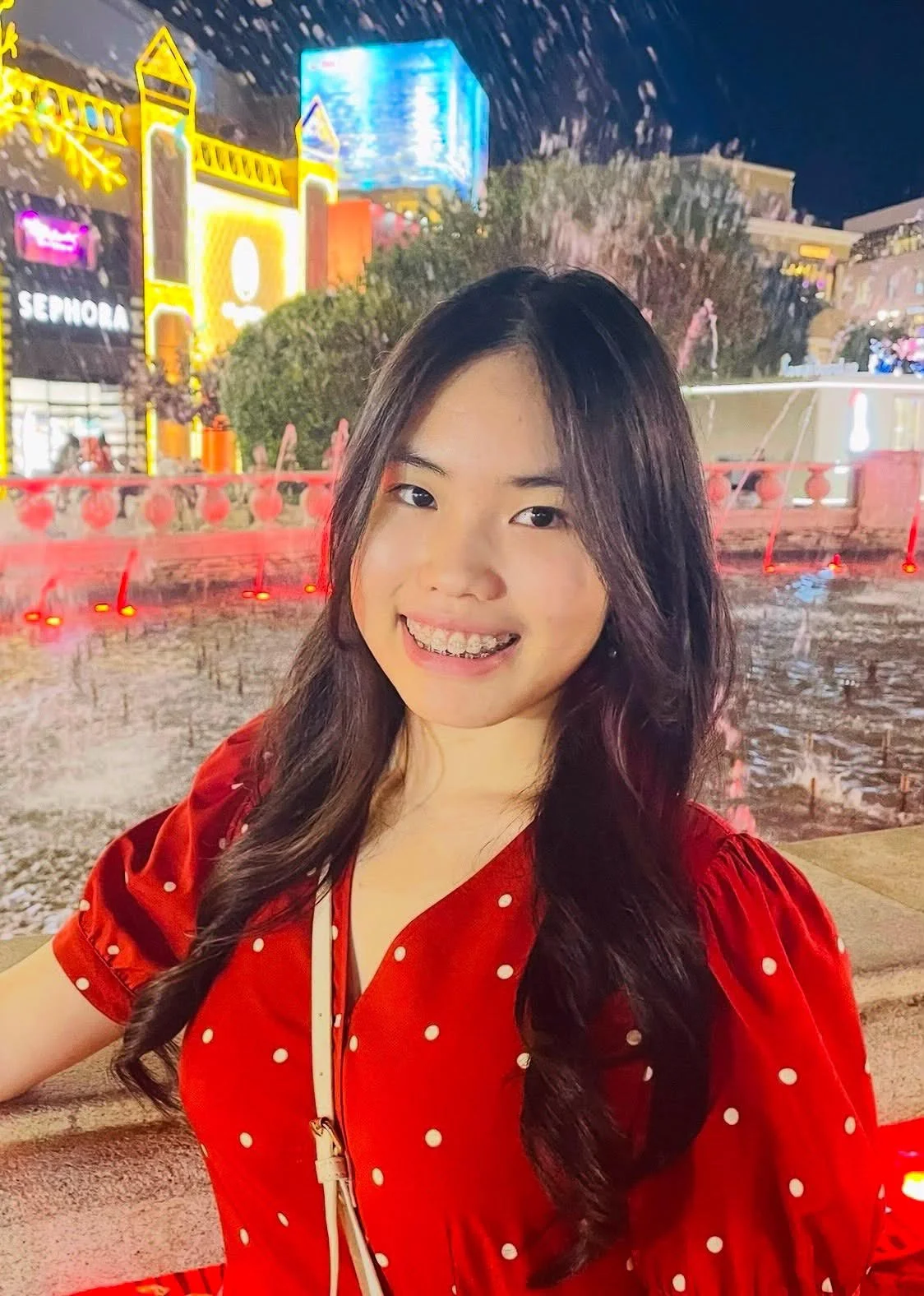 A young woman with long dark hair smiling in front of a brightly lit urban night scene with colorful building facades and a fountain.
