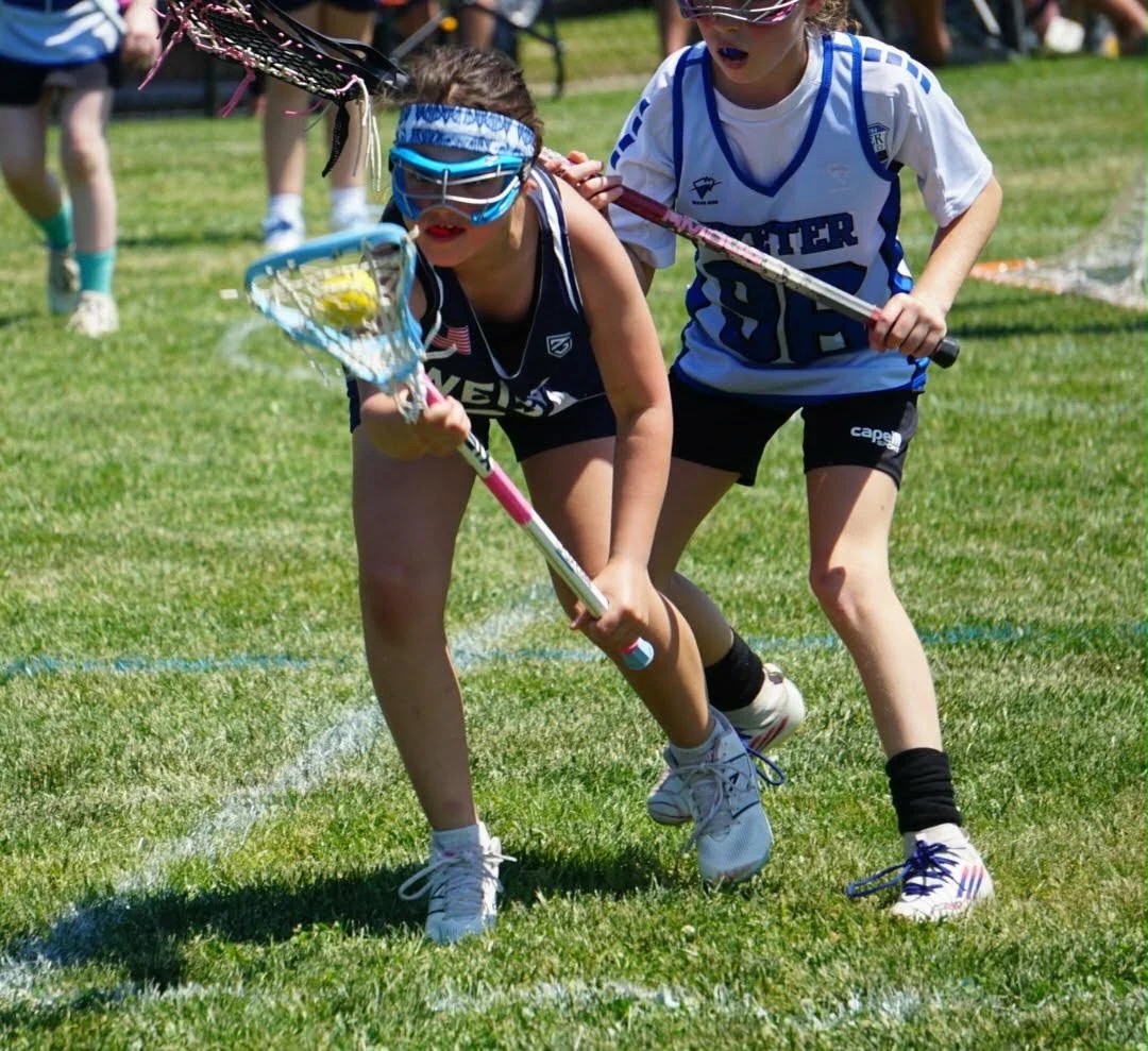 Two young girls playing lacrosse outdoors on a grassy field. One girl is holding a lacrosse stick with a yellow ball, wearing sunglasses, a headband, and sports gear, while the other girl is trying to defend, also with a lacrosse stick, wearing sport