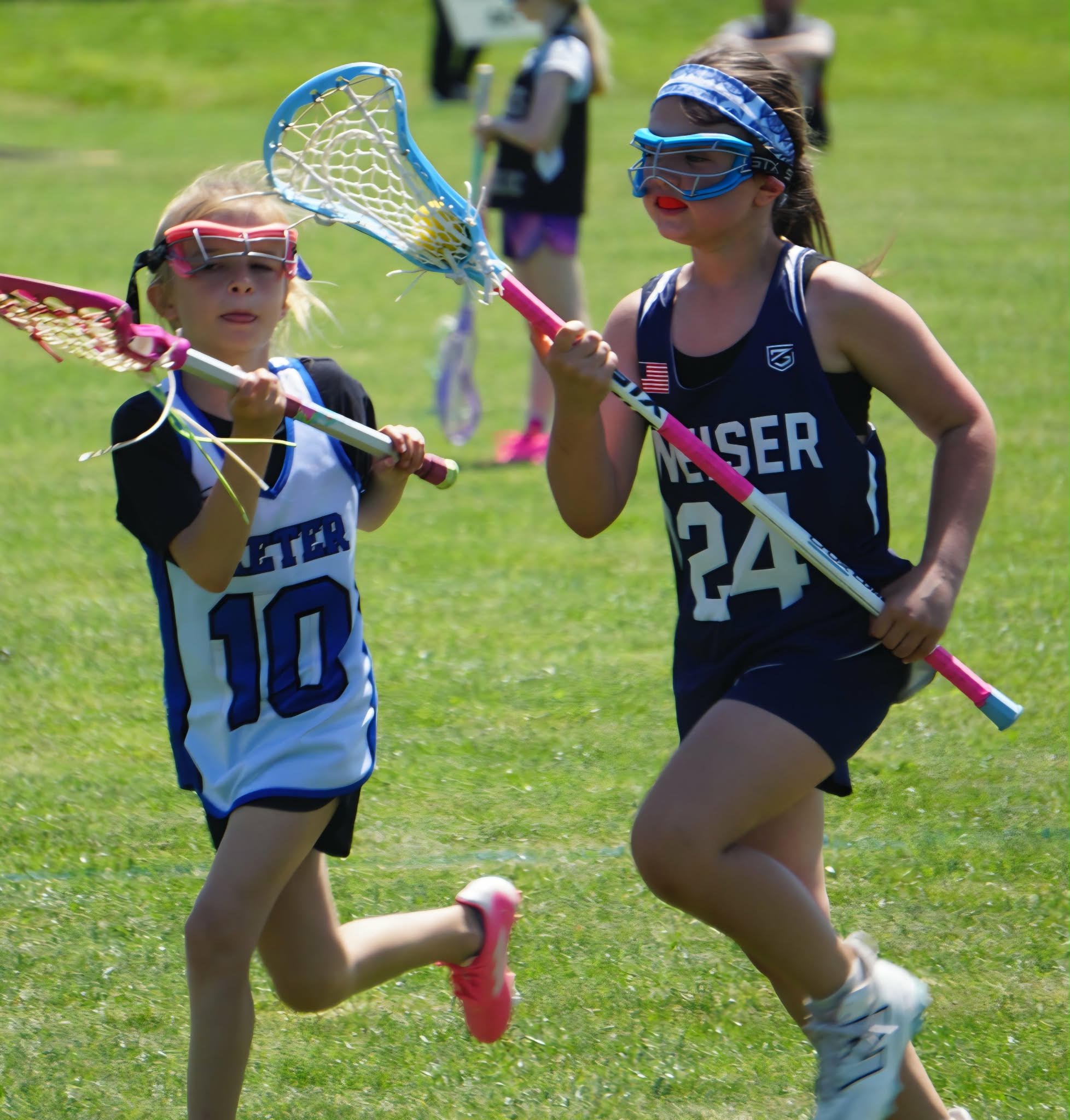 Two young girls playing lacrosse on a grassy field, wearing sports uniforms and goggles, with other children in the background.