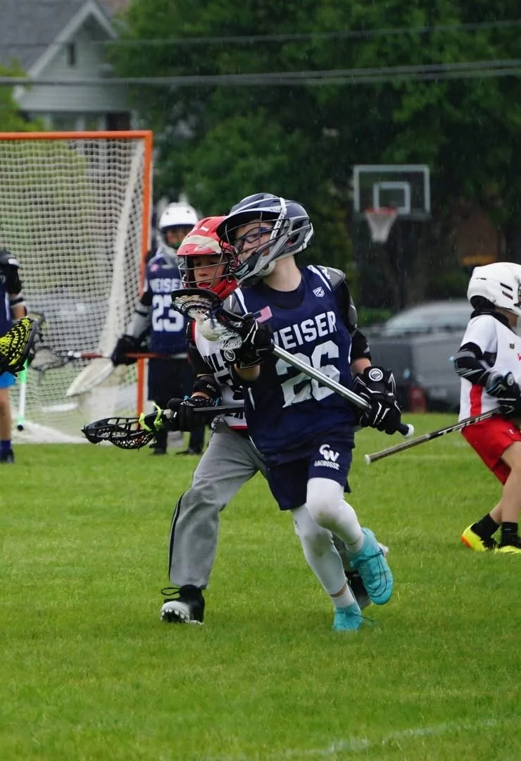 Lacrosse players in gear, including helmets and gloves, competing on a grassy field during daytime; one player in a blue uniform with the number 26 is forefront, with other players and sports equipment visible in the background.