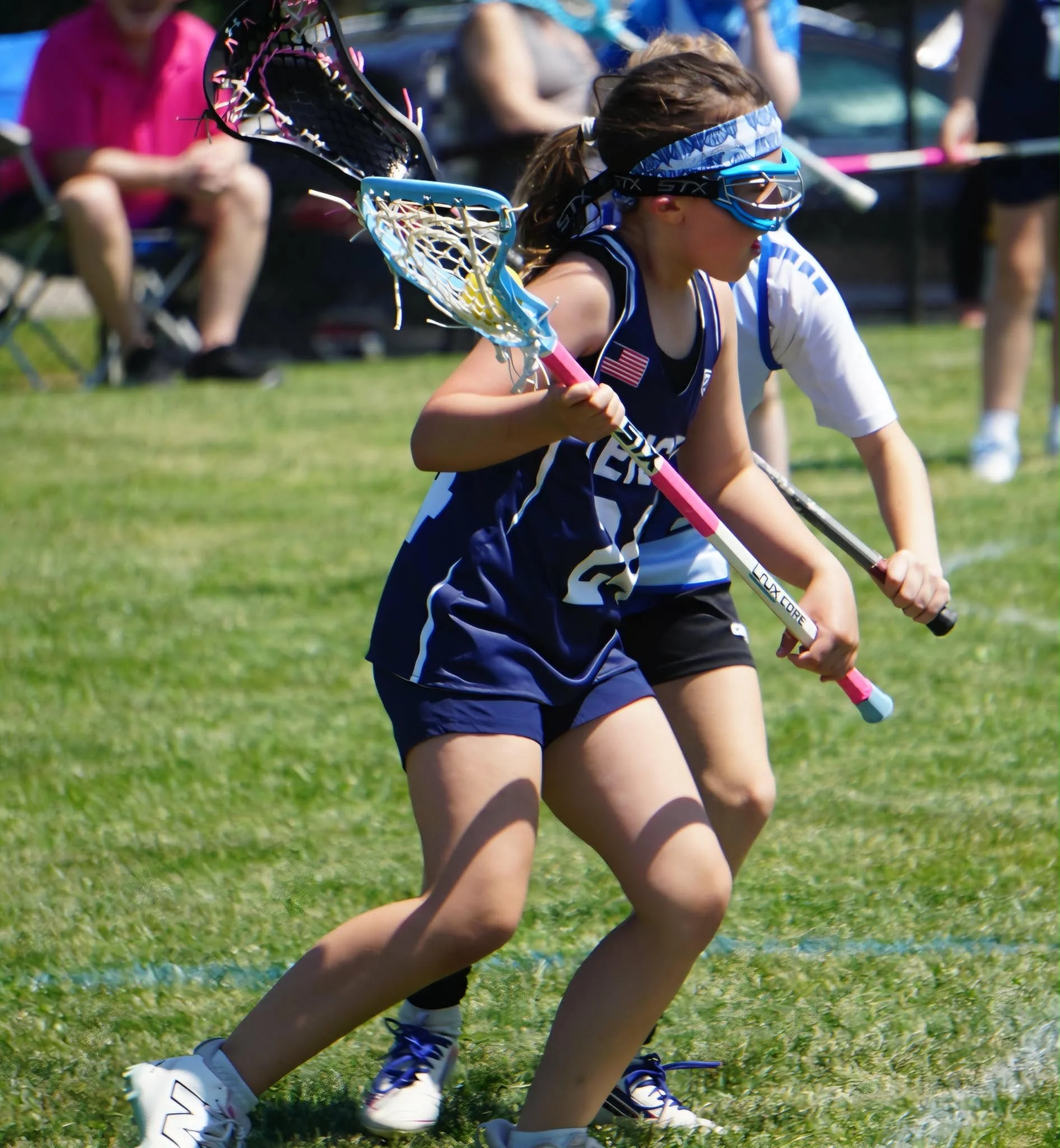 A female lacrosse player in a blue and white uniform crouches on the grass holding a lacrosse stick, wearing protective goggles and a headband, with other players and spectators in the background.