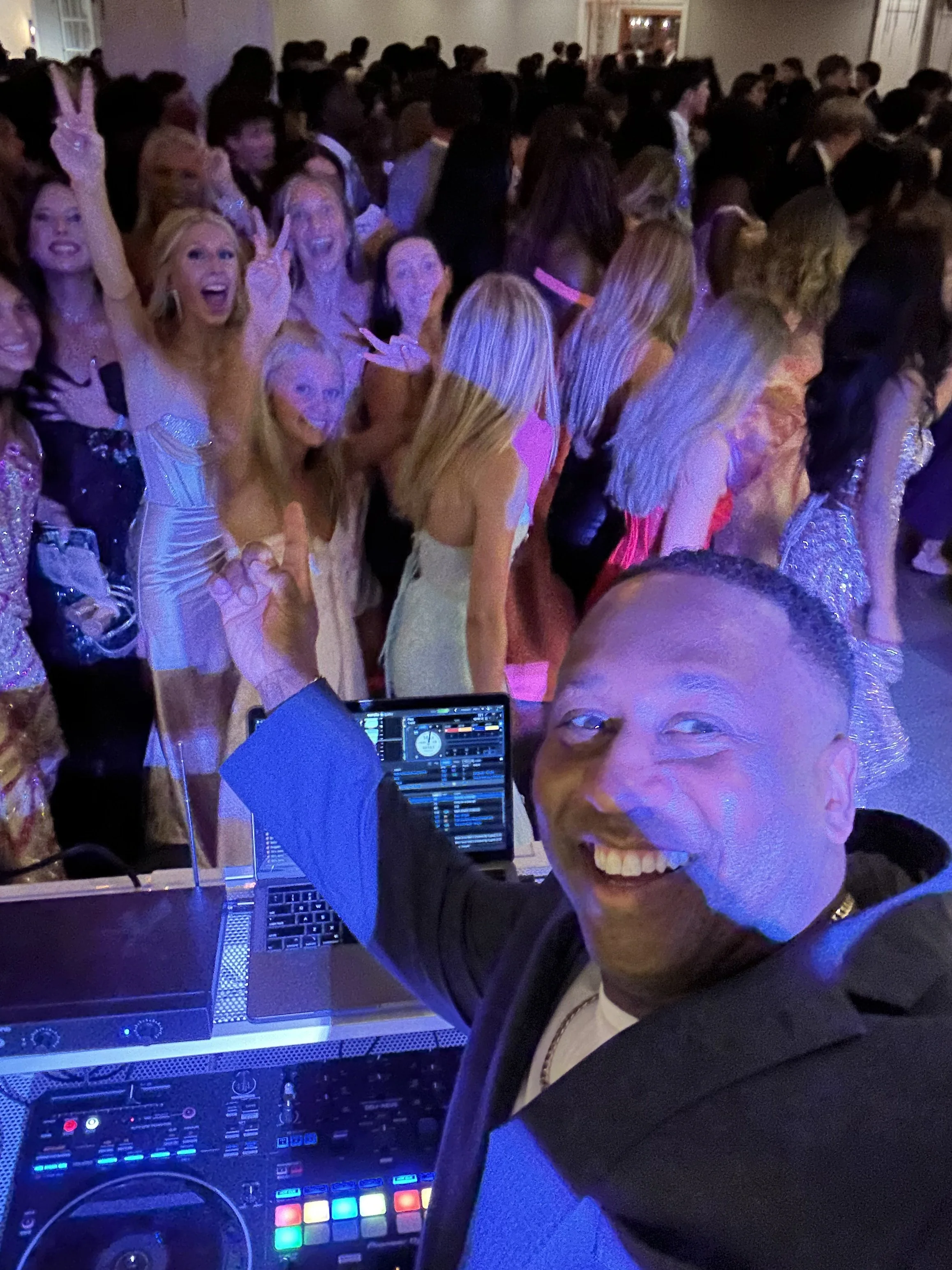 A man smiling and taking a selfie at a crowded party with many women in evening gowns dancing in the background. A DJ setup with colorful lights and a laptop is also visible.