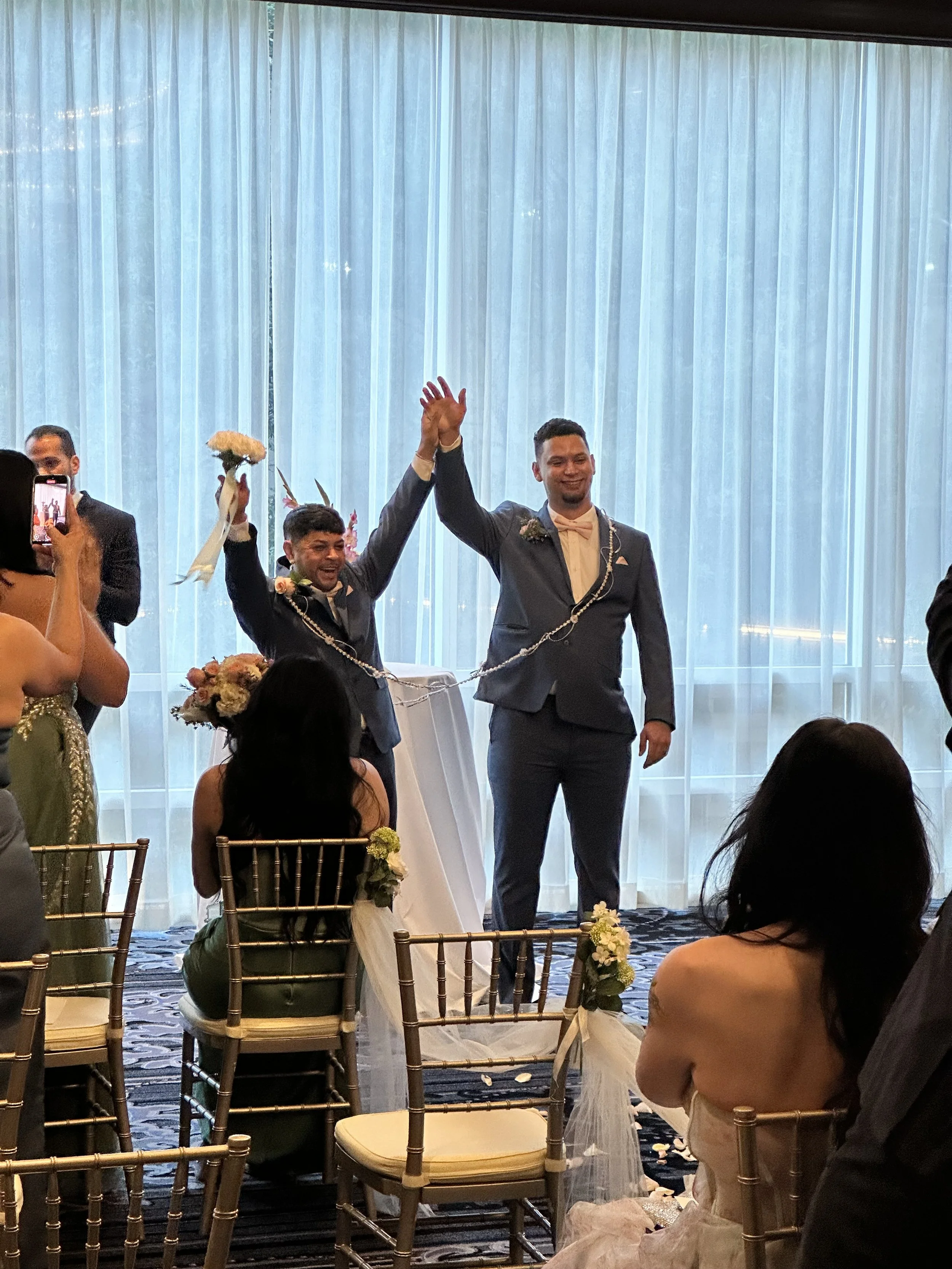 Two grooms in suits holding hands in a wedding ceremony, celebrating after the vows, with guests seated around them and a blue curtain backdrop.