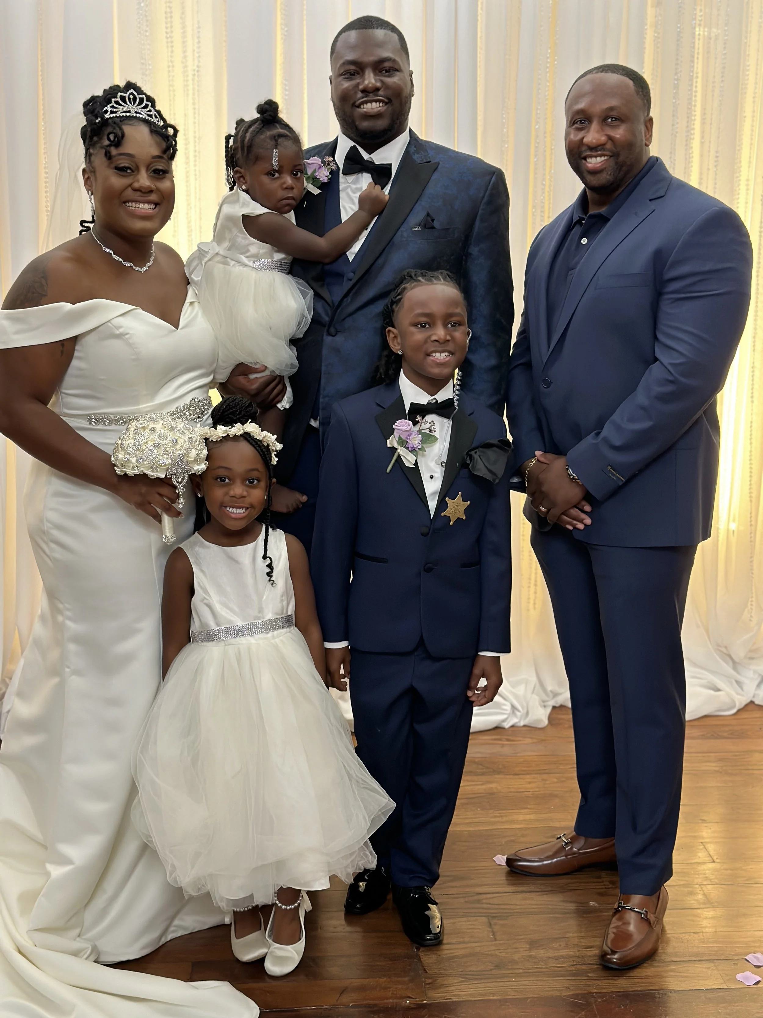 A wedding reception, dressed in formal attire, standing in front of a cream-colored curtain with string lights. The bride wears a white off-shoulder gown with a tiara and jewelry. The groom wears a blue suit with a black bow tie. Two young girls wear white dresses with flower embellishments, and an older boy wears a navy tuxedo with a sheriff badge. A man in a blue suit stands beside them.