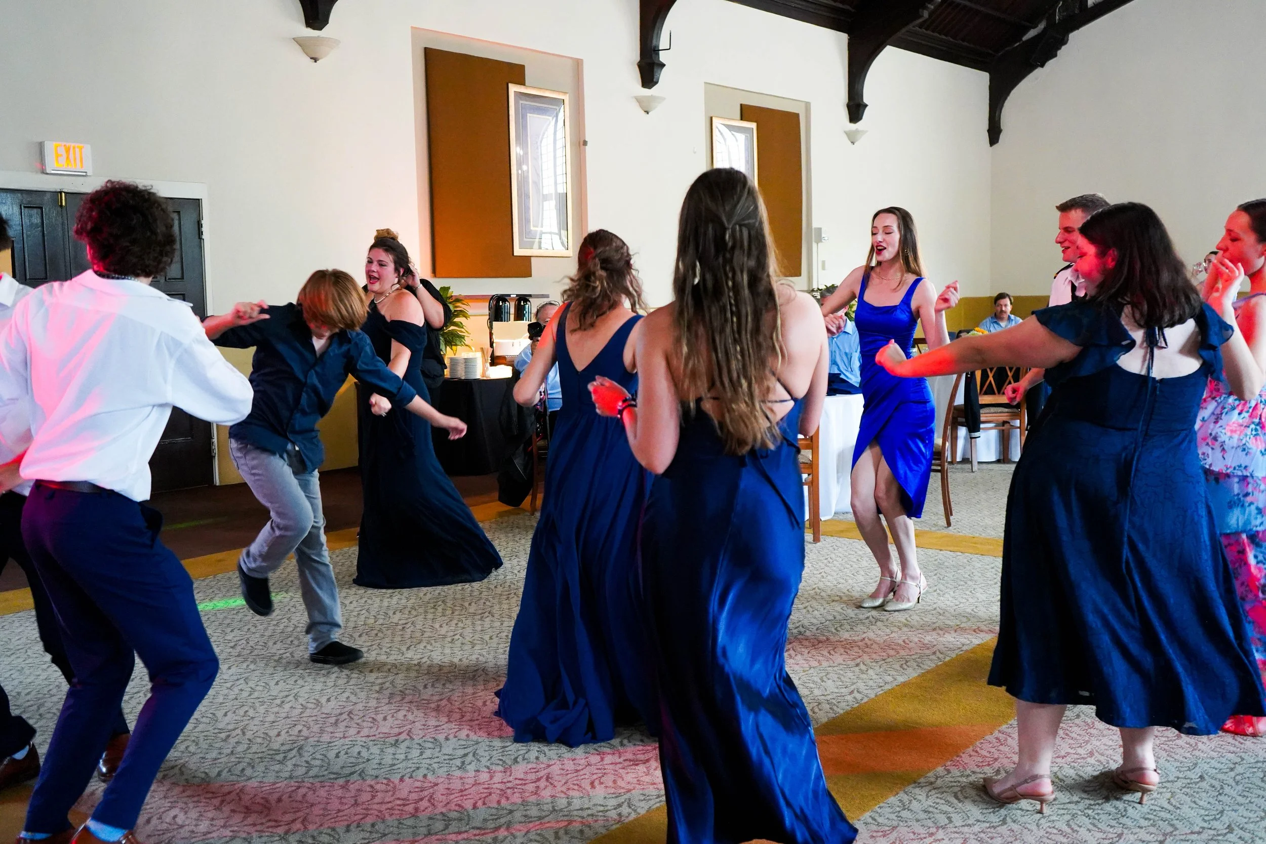 People dancing at a celebration or party in a banquet hall, with women in blue dresses and men in formal attire.