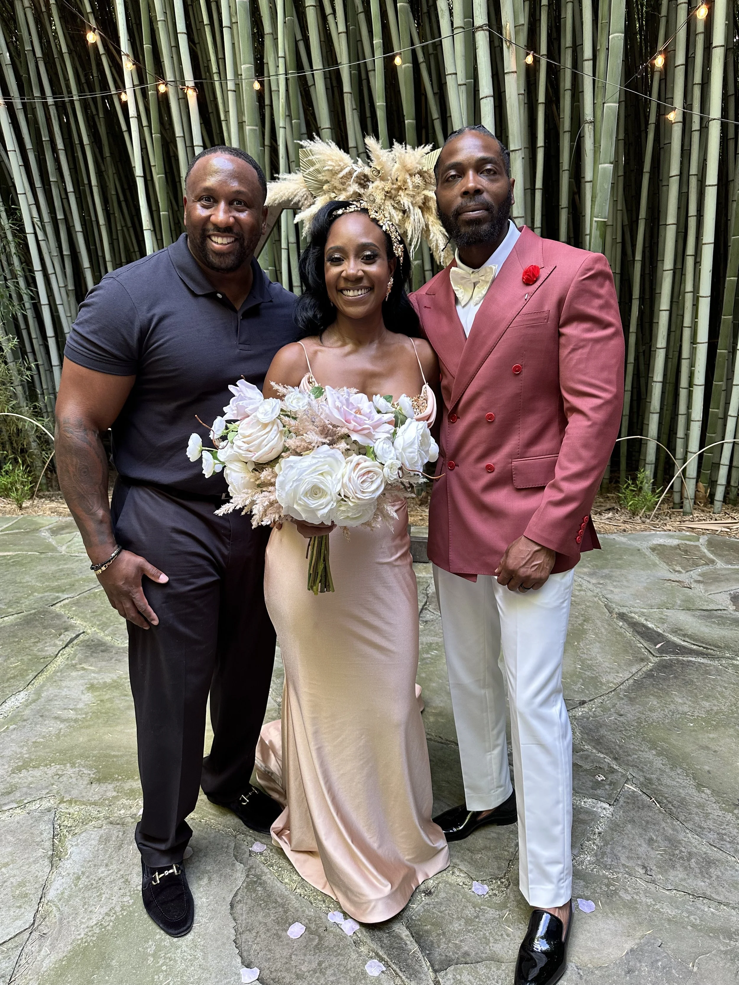 A woman in a light pink satin gown holding a bouquet of white roses, flanked by two men in formal attire, one in a black shirt and the other in a red blazer with white pants, standing in front of a bamboo backdrop with string lights.