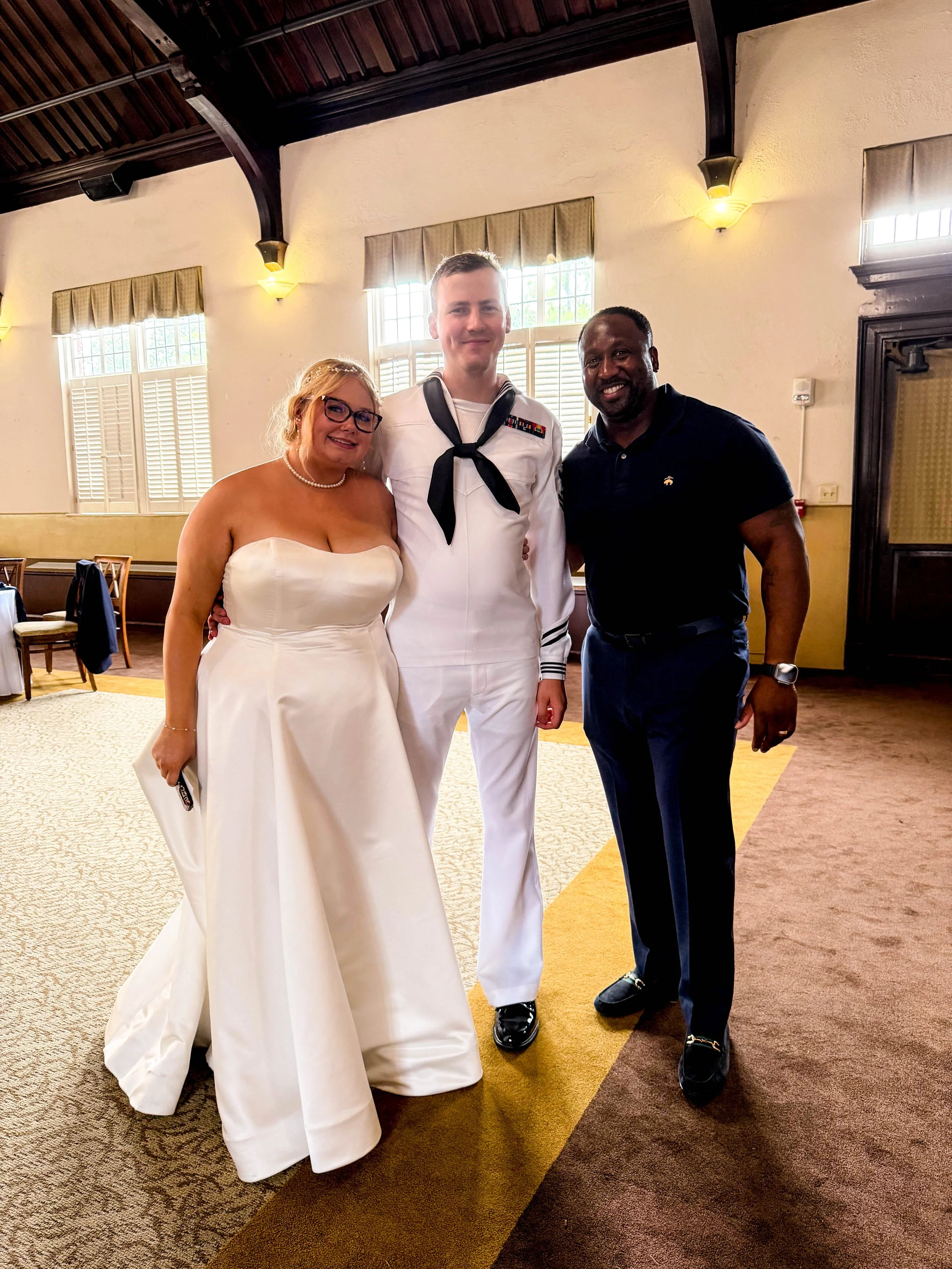 Three people standing together indoors, smiling at the camera. The woman on the left is in a strapless white wedding gown with glasses and a pearl necklace. The man in the middle is in a white military uniform with a black neckerchief and medals. The man on the right is in a black polo shirt and dark pants.