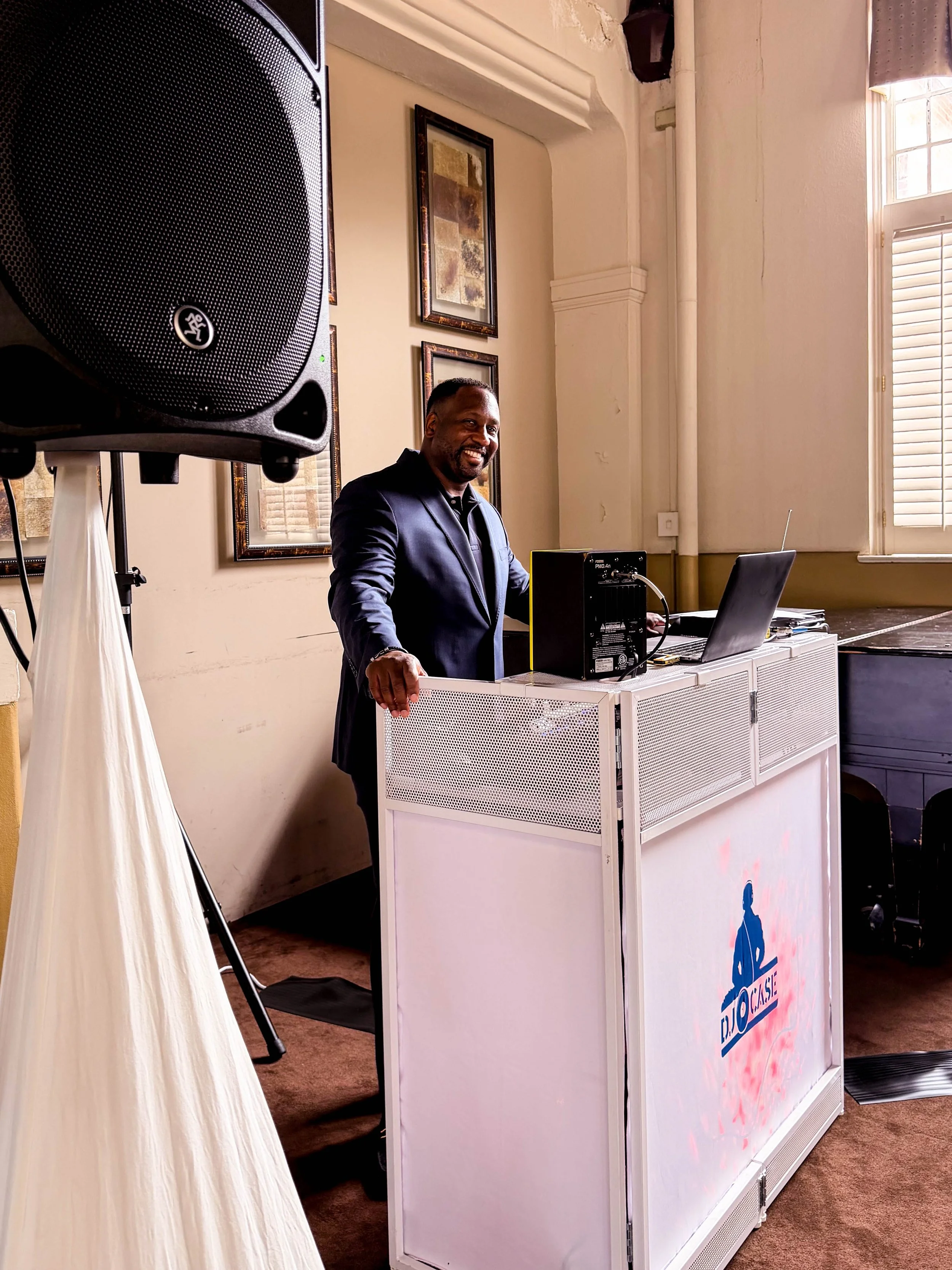 A man in a dark tuxedo standing behind a DJ booth, smiling. The booth has a logo with a silhouette of a person painting and the words 'D.ROUSE'. Large speaker on a stand in the foreground, some framed pictures on the wall, a window with blinds, and a stack of papers on the table.