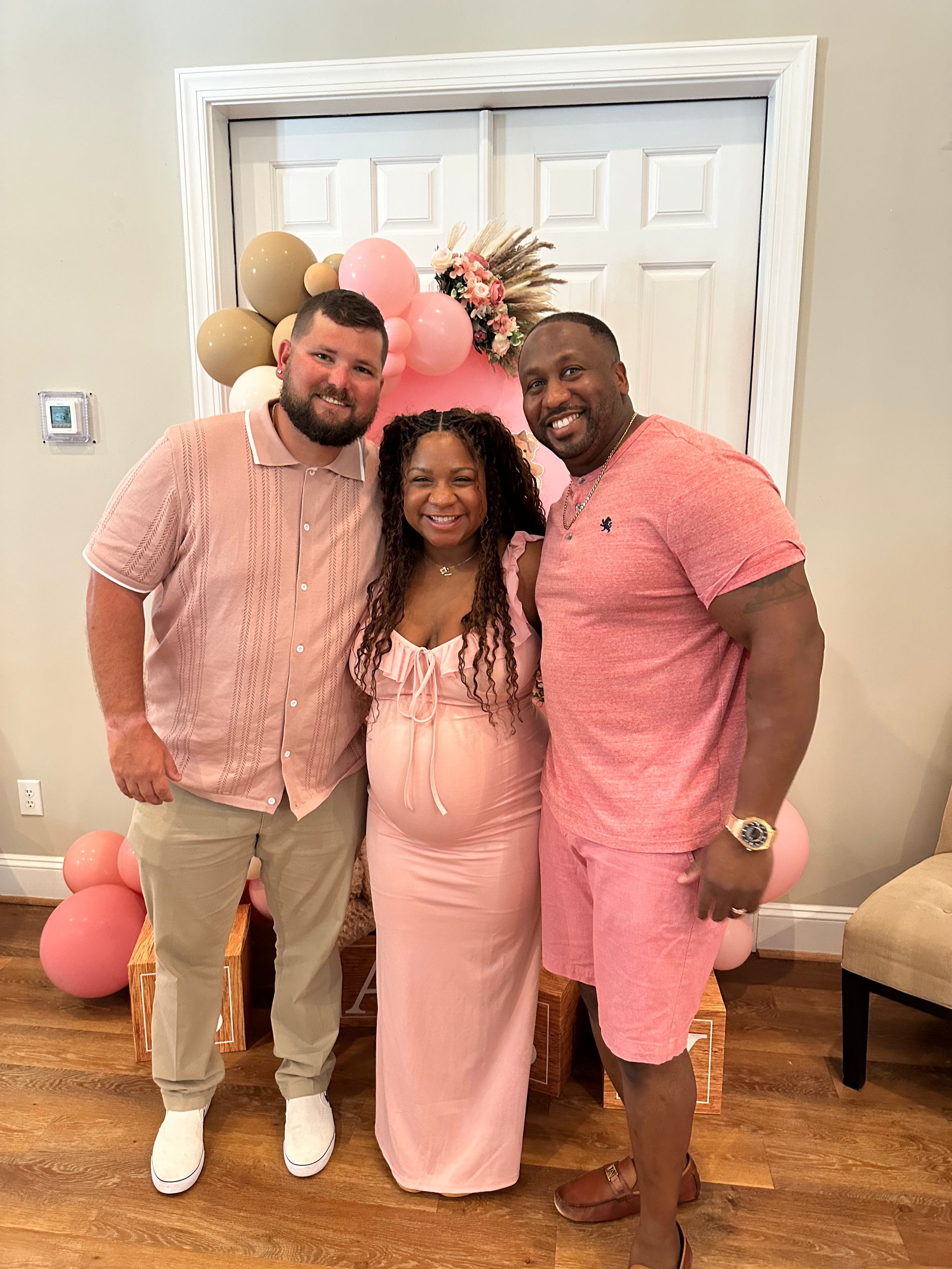 Three people, two men and one woman, smiling and posing for a photo in front of a pink balloon backdrop with floral decorations, celebrating a special occasion indoors.