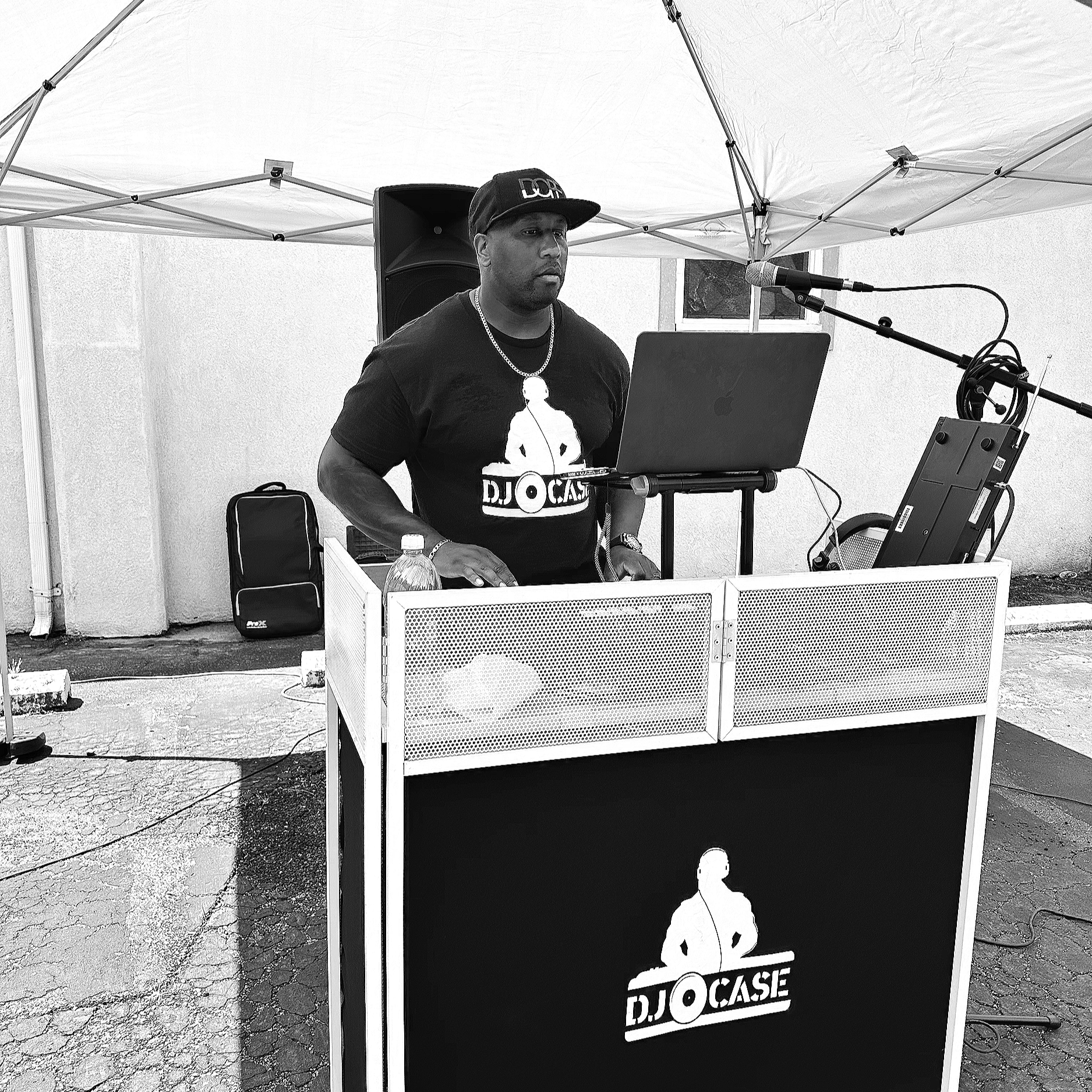A DJ at a stand under a canopy, with DJ equipment, a laptop, and a speaker, in an outdoor setting.