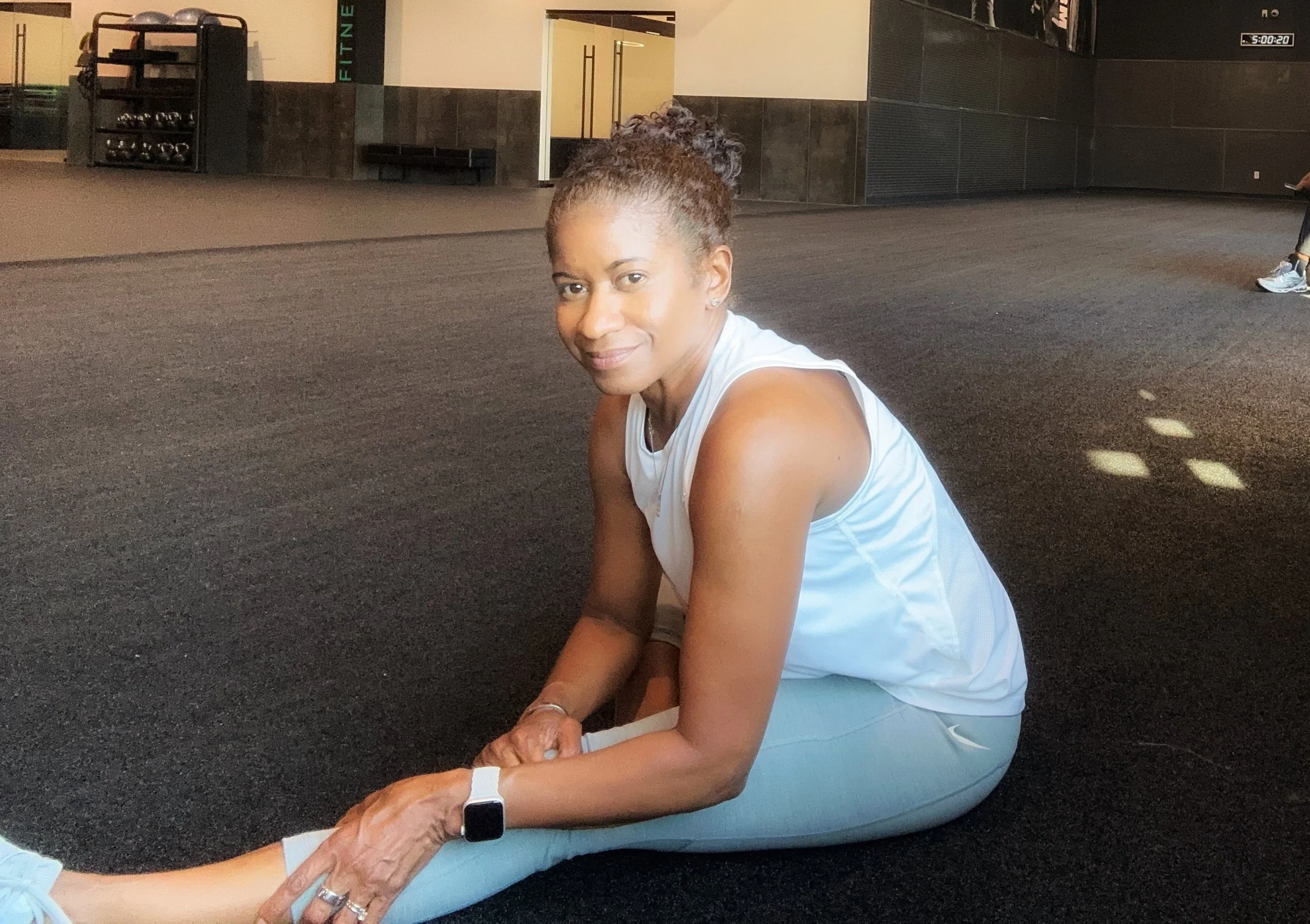 Bernadette Douglas, wellness coach and founder of Soulful Health Coach on the floor in a gym stretching, wearing an athletic tank top and light blue leggings, with fitness equipment and a black wall in the background.