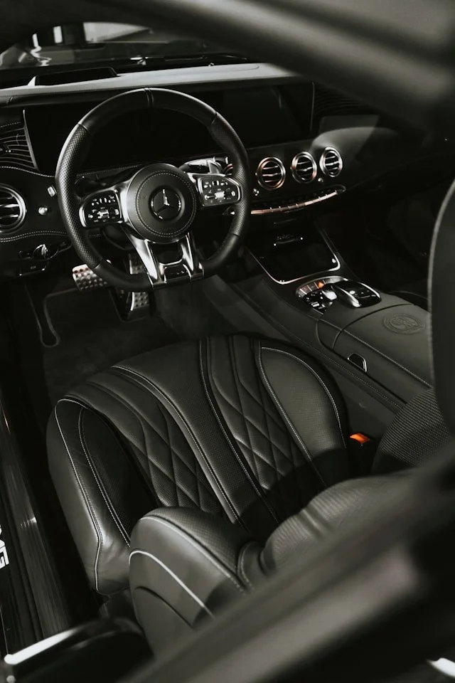 Interior of a luxury car with a black leather seat featuring quilted stitching, a multifunction steering wheel with a Mercedes-Benz emblem, three circular air vents on the dashboard, and a center console with a control knob.