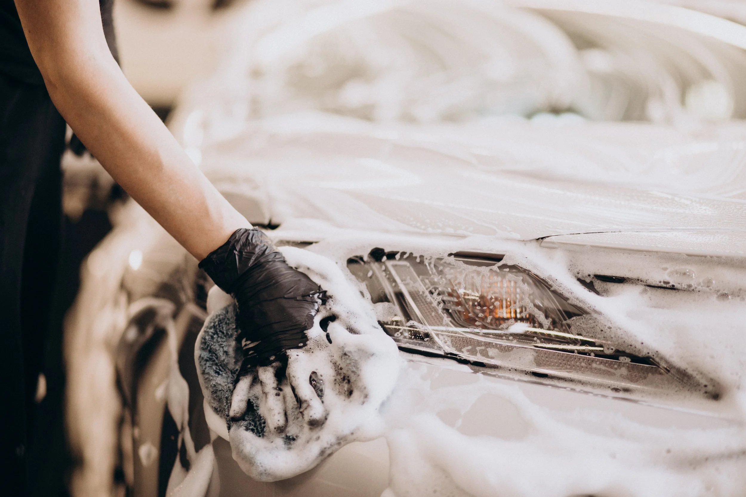 A person wearing black gloves washing a car with soap and water, creating foam on the vehicle.