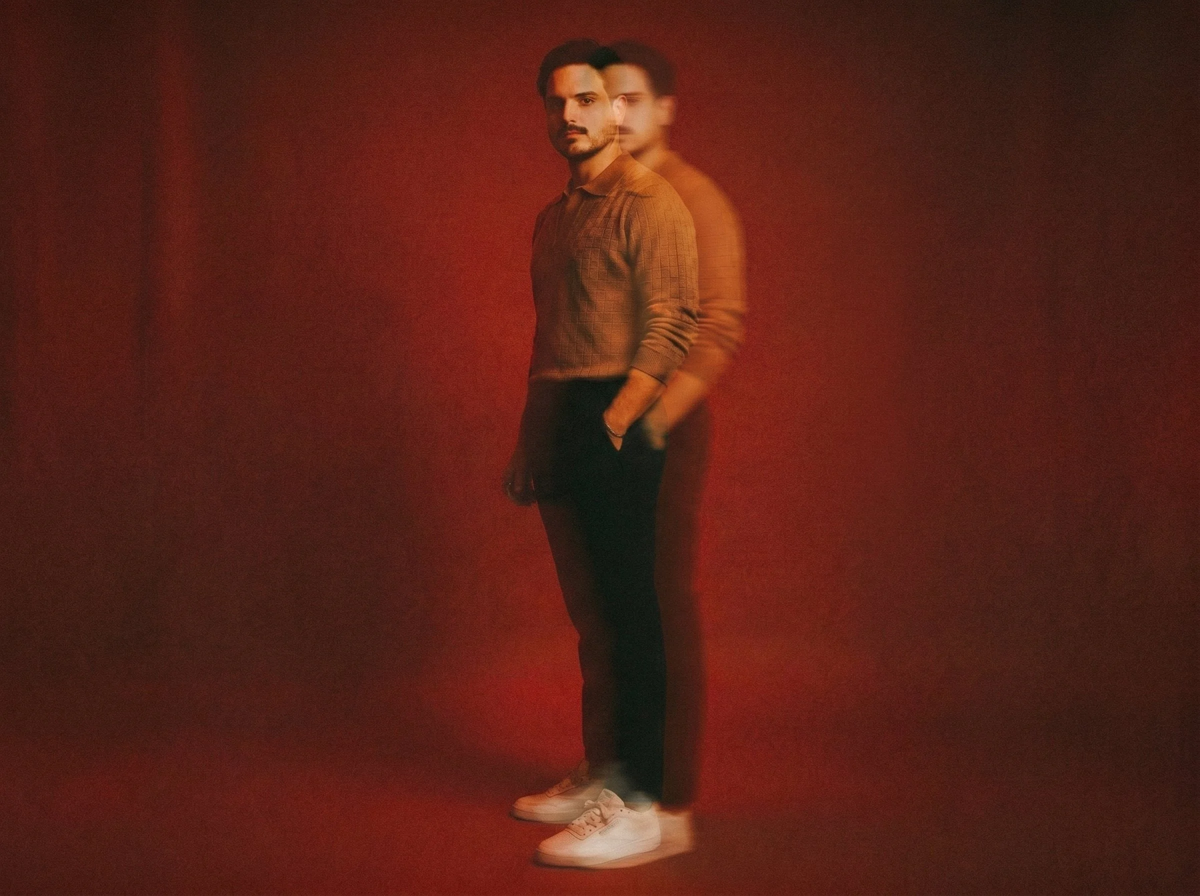 A man standing against a dark red background with a layered, blurry effect. He has dark hair, a beard, and is wearing a brown checkered shirt, black pants, and white sneakers, with his hands in his pockets.