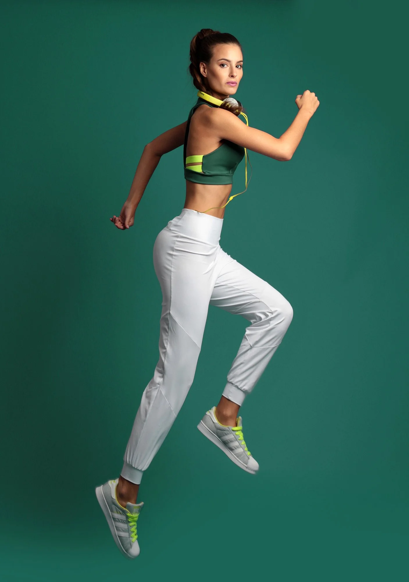 A woman in athletic clothing running or jumping mid-air against a green background, wearing a green sports top, white pants, and white sneakers with yellow accents.