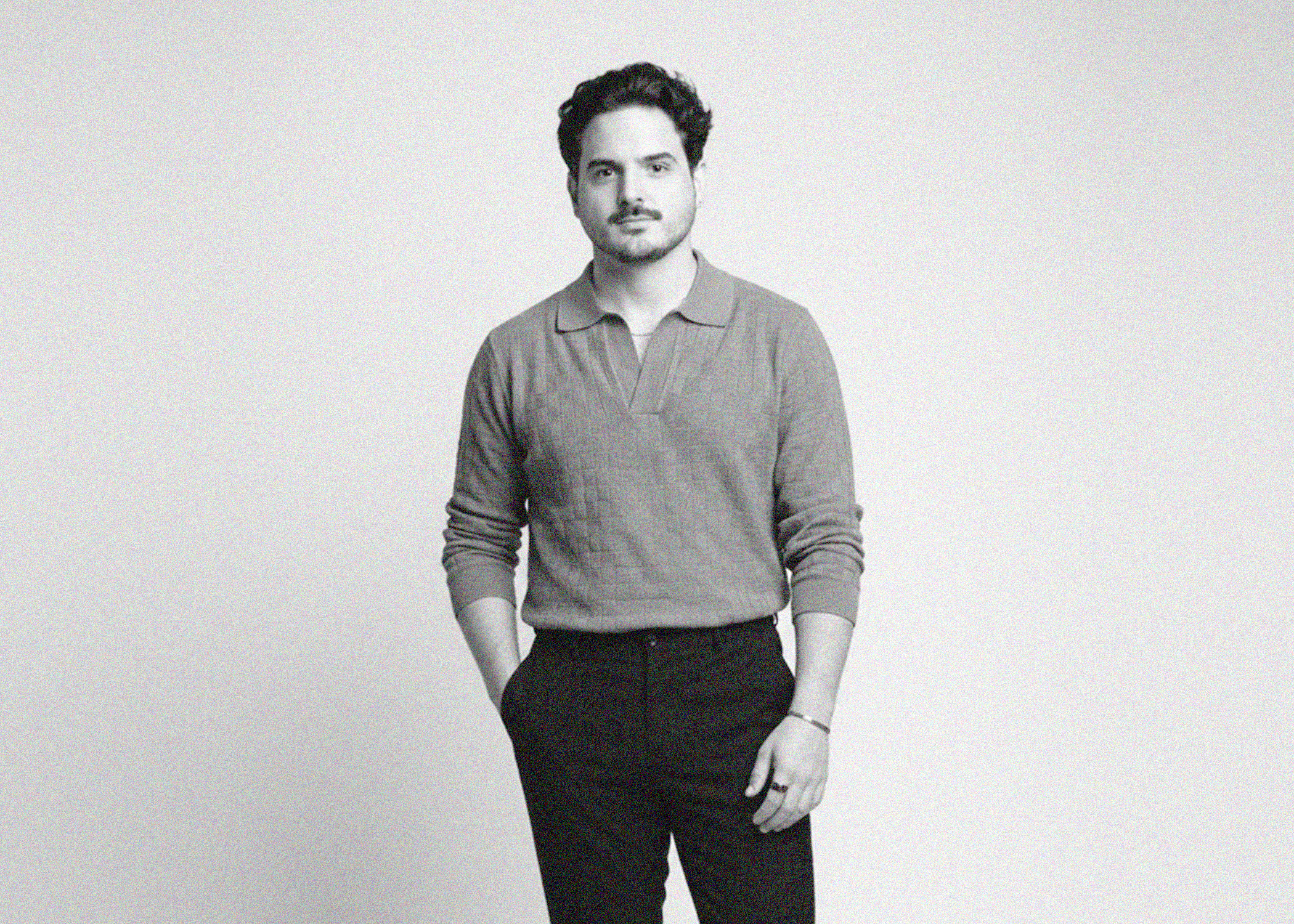 Black and white photo of a young man with dark hair and a mustache, wearing a v-neck sweater with a collared shirt underneath, standing against a plain light-colored wall.