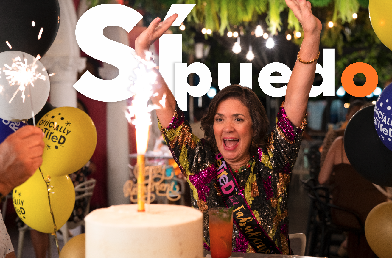 A woman celebrating her birthday at a party, wearing a colorful sequin top and a "Graduate" sash, with a birthday cake, balloons, and sparklers around her.