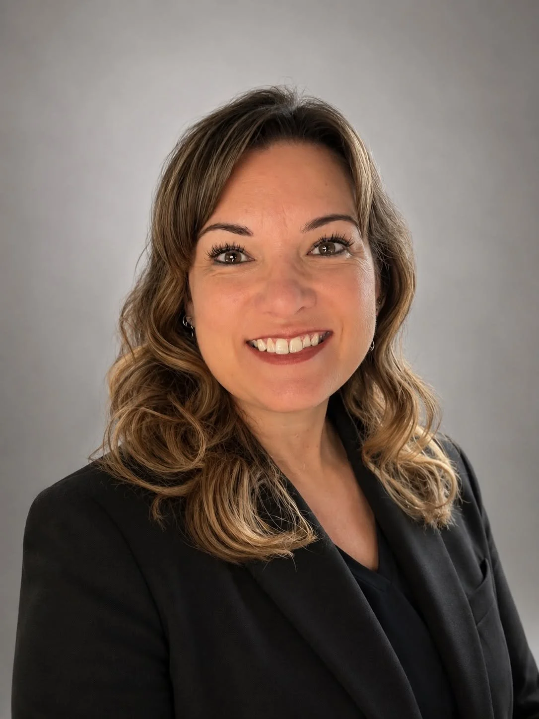 A woman with wavy light brown hair and wearing a black blazer, smiling against a plain gray background.