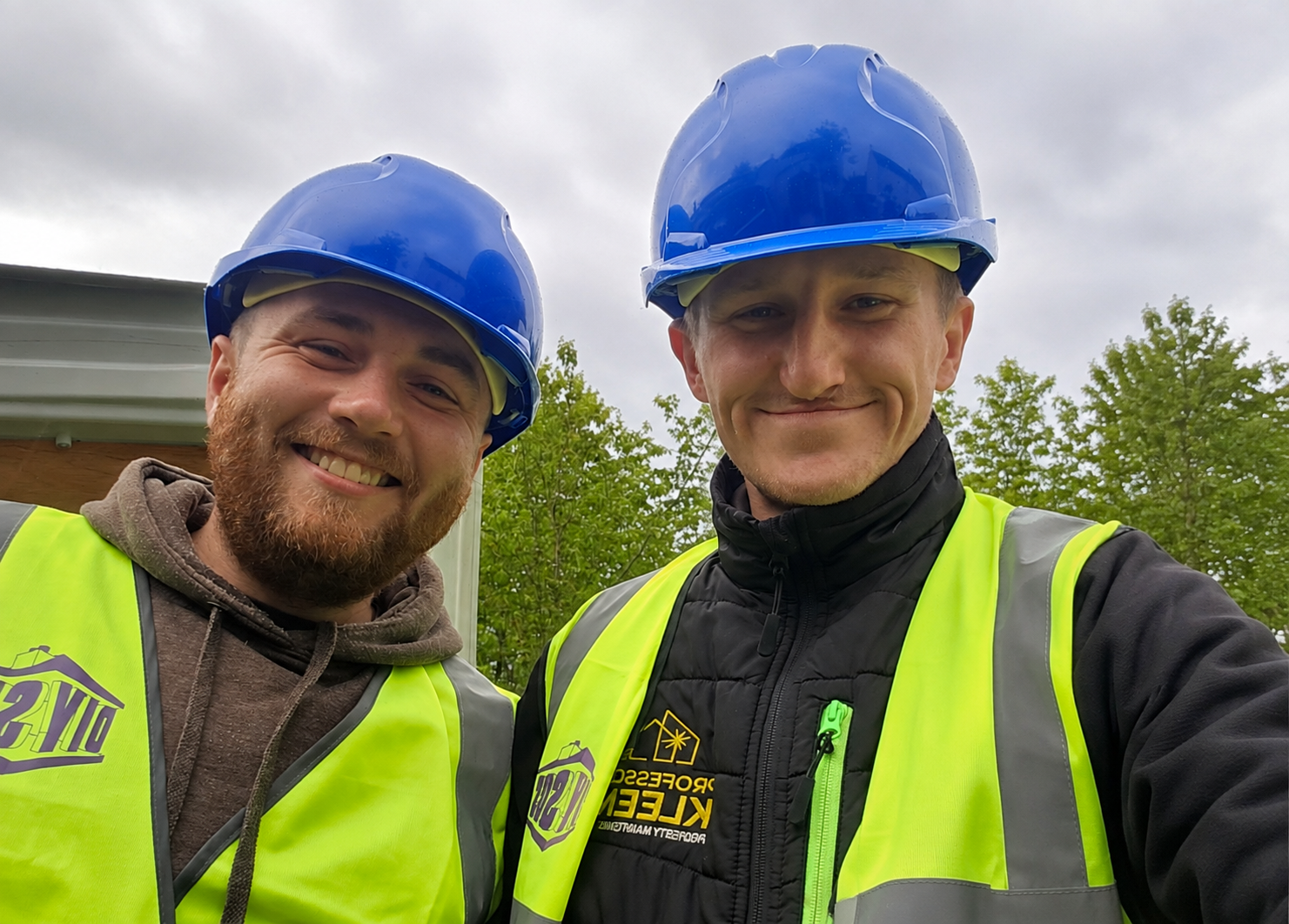 Two construction workers wearing blue safety helmets and bright yellow safety vests, smiling for a selfie outside with trees and a cloudy sky in the background.