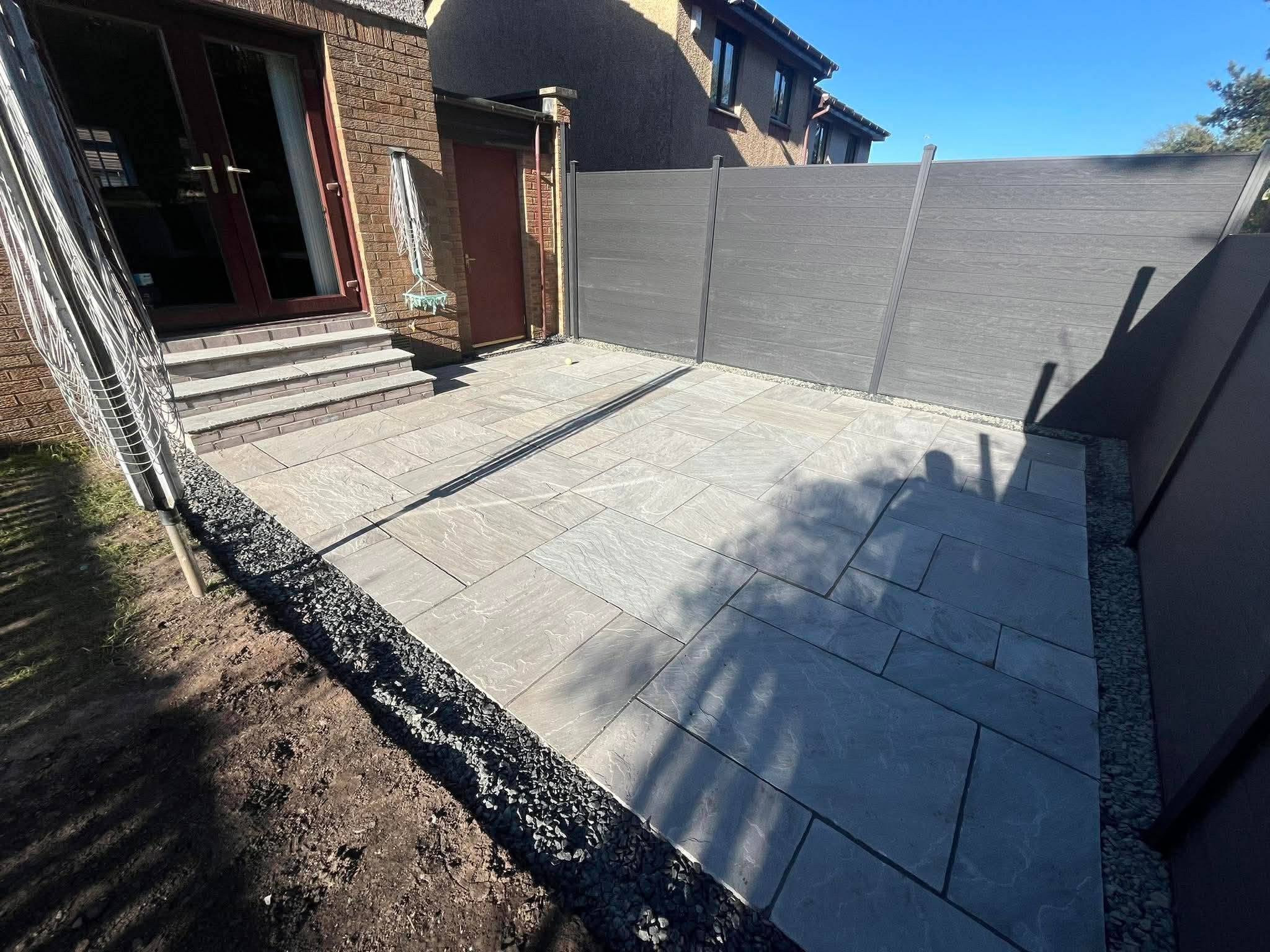 A backyard patio with light gray tiles, a gray fence on the right, a brick house with a glass door and stairs on the left, and a small door near the stairs. There is a clothesline pole and a white round hanging basket near the stairs. The sky is clear and blue.
