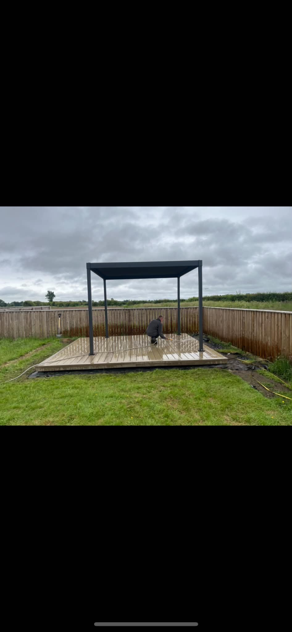 A man working on a wooden deck with a black metal frame structure, surrounded by a wooden fence and green grass, overcast sky in the background.