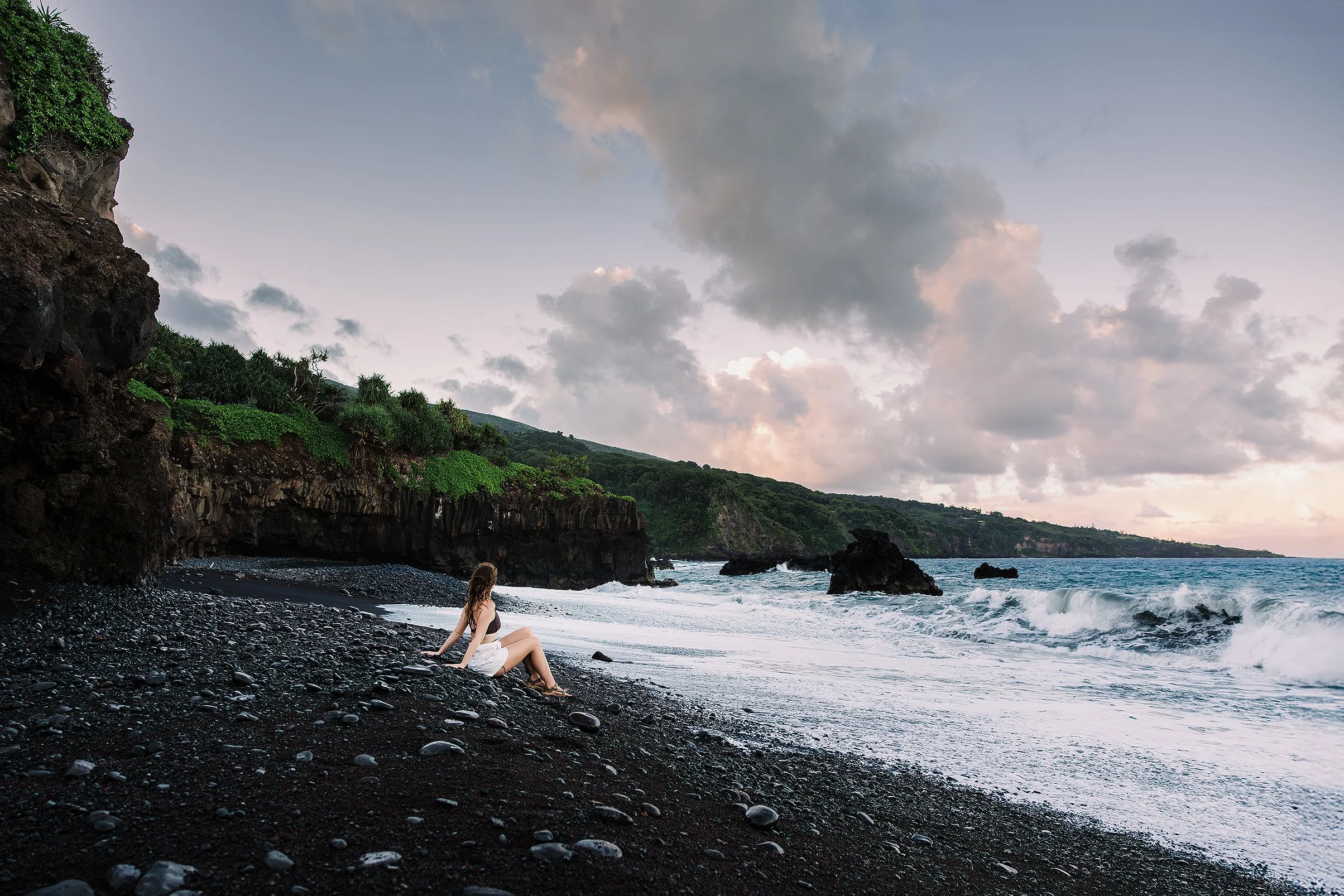 A person sitting on a pebbled black beach near the ocean with rocky cliffs and green vegetation in the background, and a cloudy sky overhead.