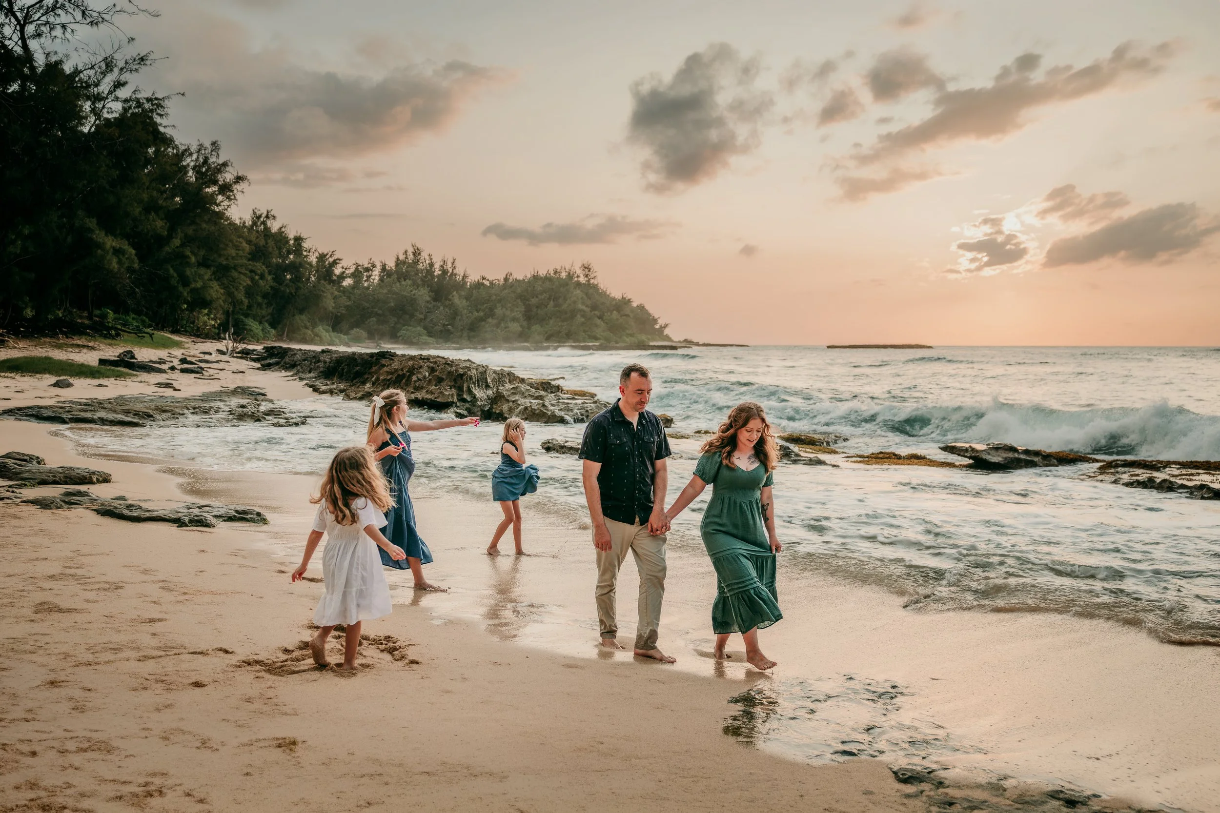 A group of people, including adults and children, walking hand in hand along the beach at sunset, with the ocean waves and cloudy sky in the background.