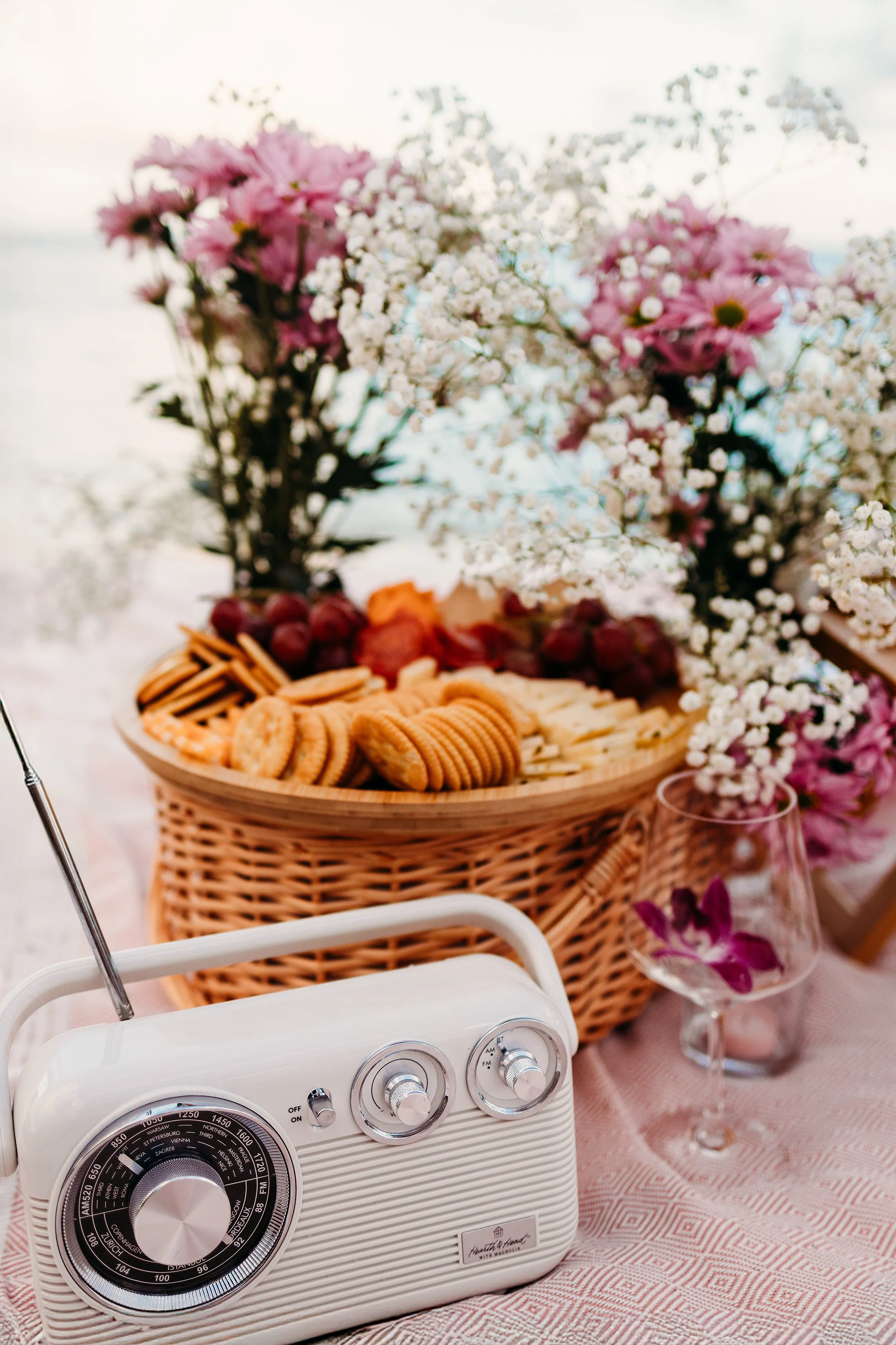 A vintage white radio with a black dial, placed on a table with a pink tablecloth, in front of a wicker basket filled with pink flowers and white baby's breath. There is a glass of purple flowers to the right of the basket.