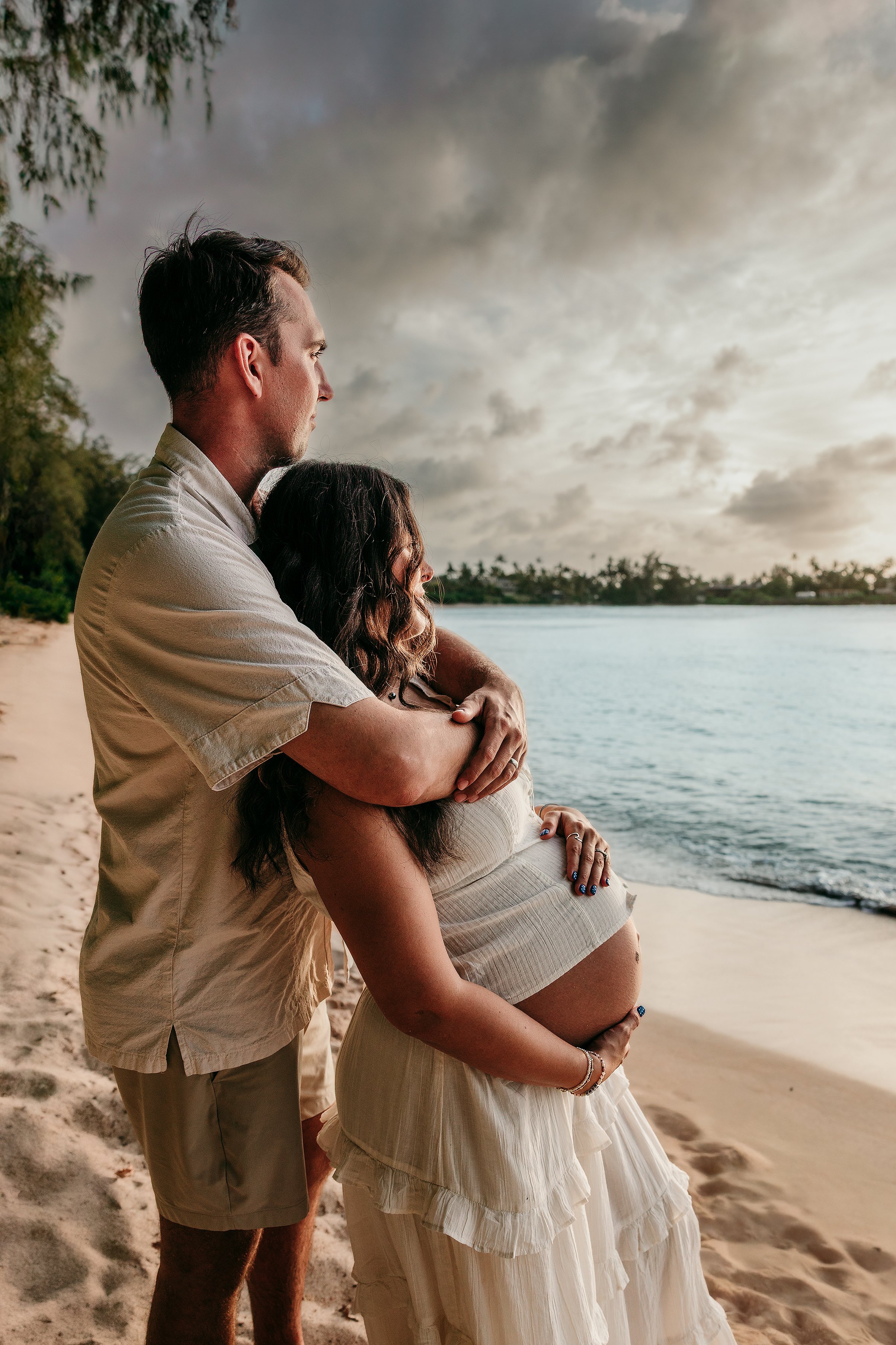 A couple stands on the beach, with the woman resting her pregnant belly as the man embraces her from behind, both facing the water with a cloudy sky above.