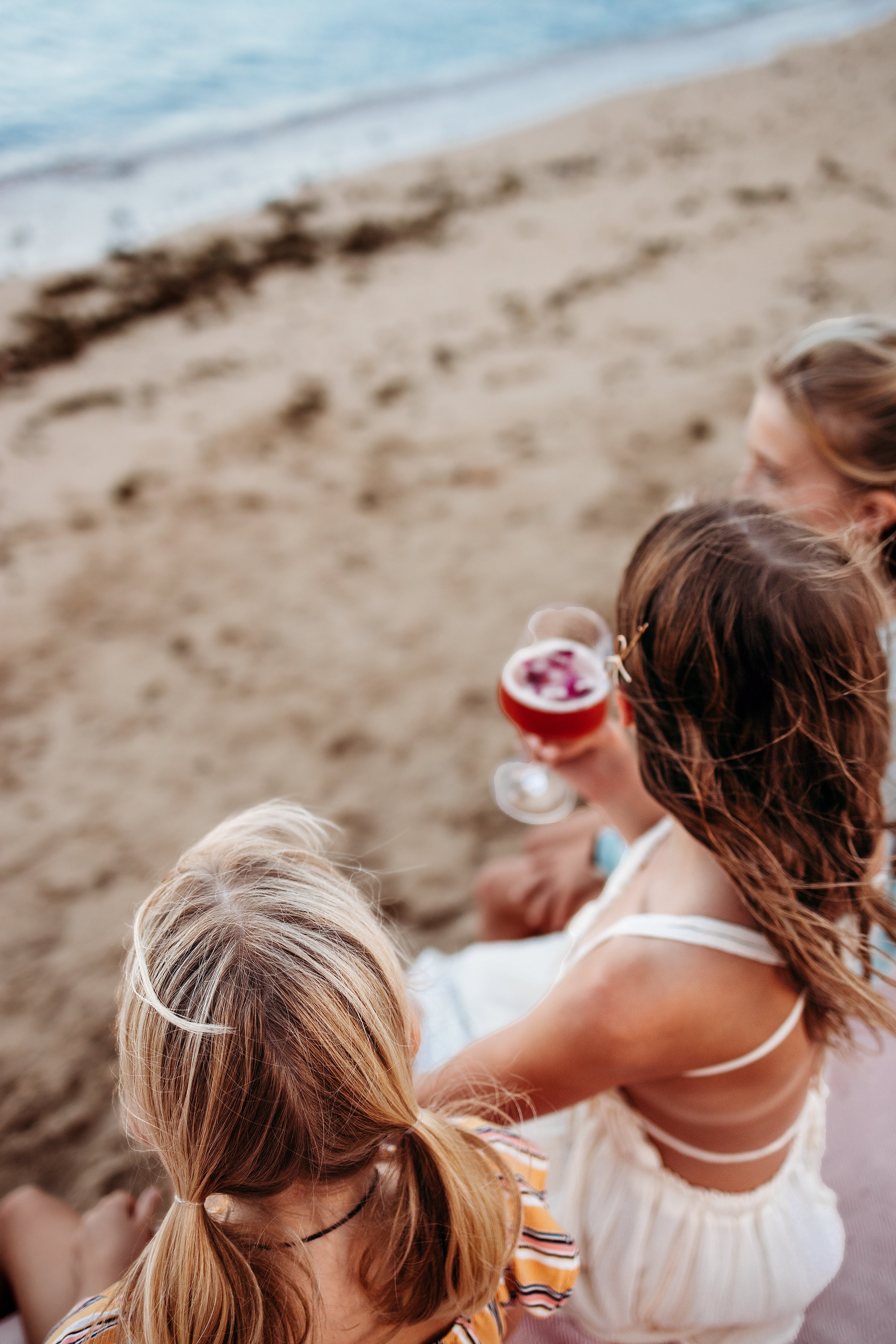Two women with long hair sitting on a sandy beach, holding drinks, with ocean waves in the background.