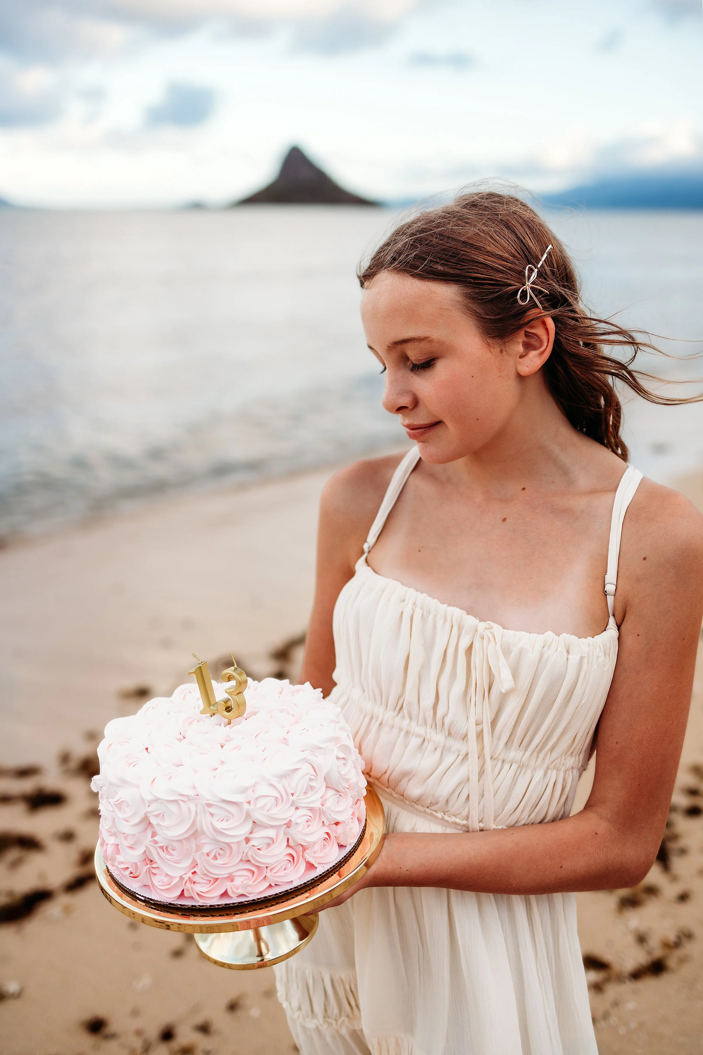 A young woman in a white dress holding a pink and white rosette cake with gold birthday candles shaped as 13 on top, on a beach with a mountain in the background.