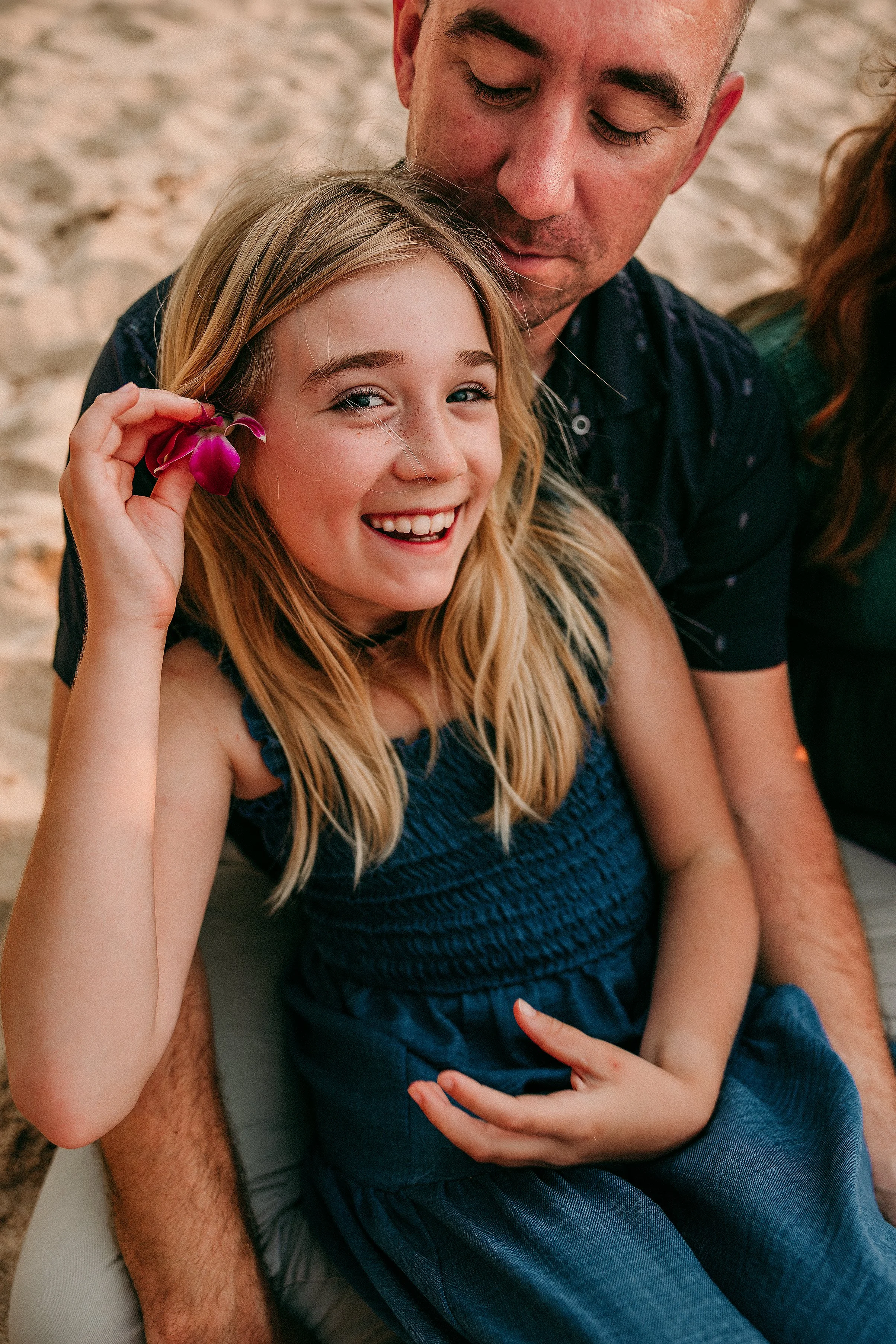 A young girl with long blonde hair, wearing a blue dress, smiling and holding a purple flower near her ear, while sitting on a person's lap on the beach. The person is an adult male with light skin and dark hair, who is gently resting his face near t