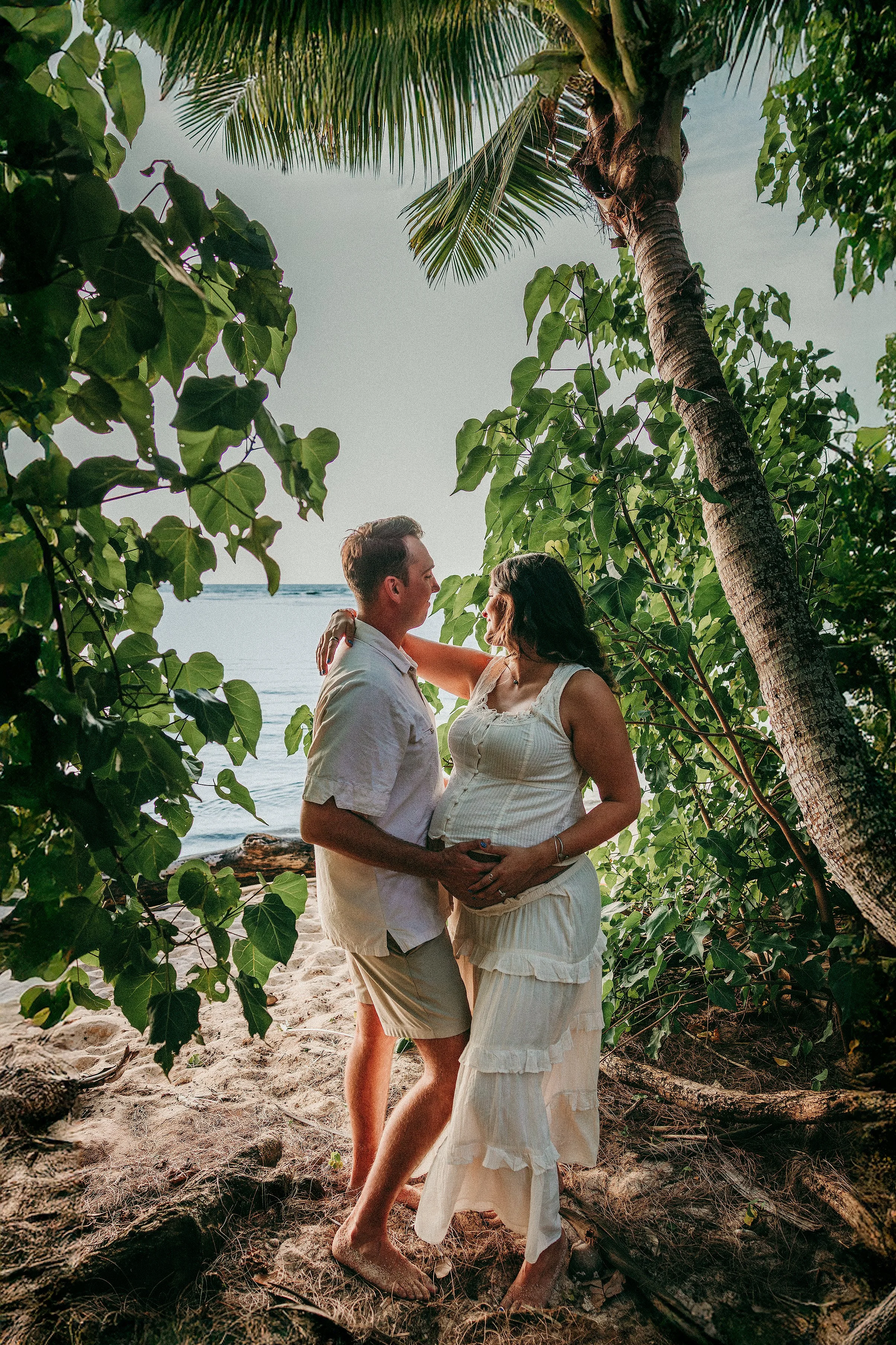 A couple standing close together on a beach, surrounded by lush green foliage and palm trees, with the ocean in the background. The woman is pregnant and wearing a white dress, and the man is wearing a light-colored shirt and shorts.