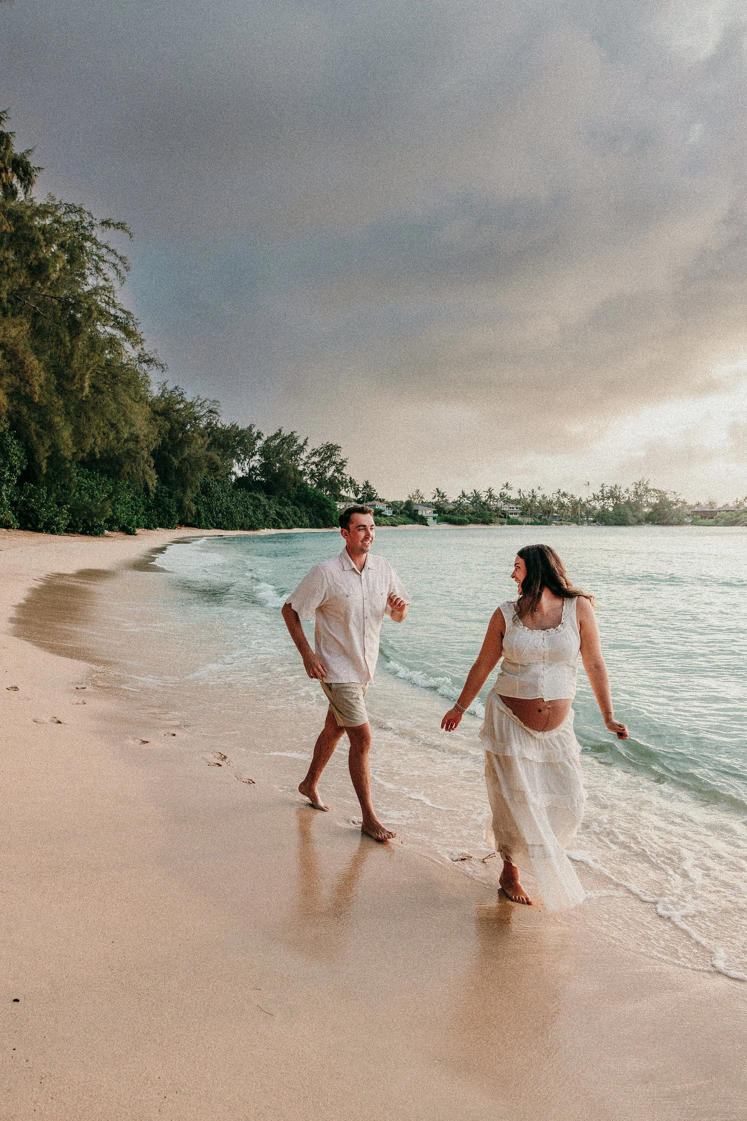 A couple walking along a beach during sunset, with the woman visibly pregnant and both smiling and enjoying the moment.