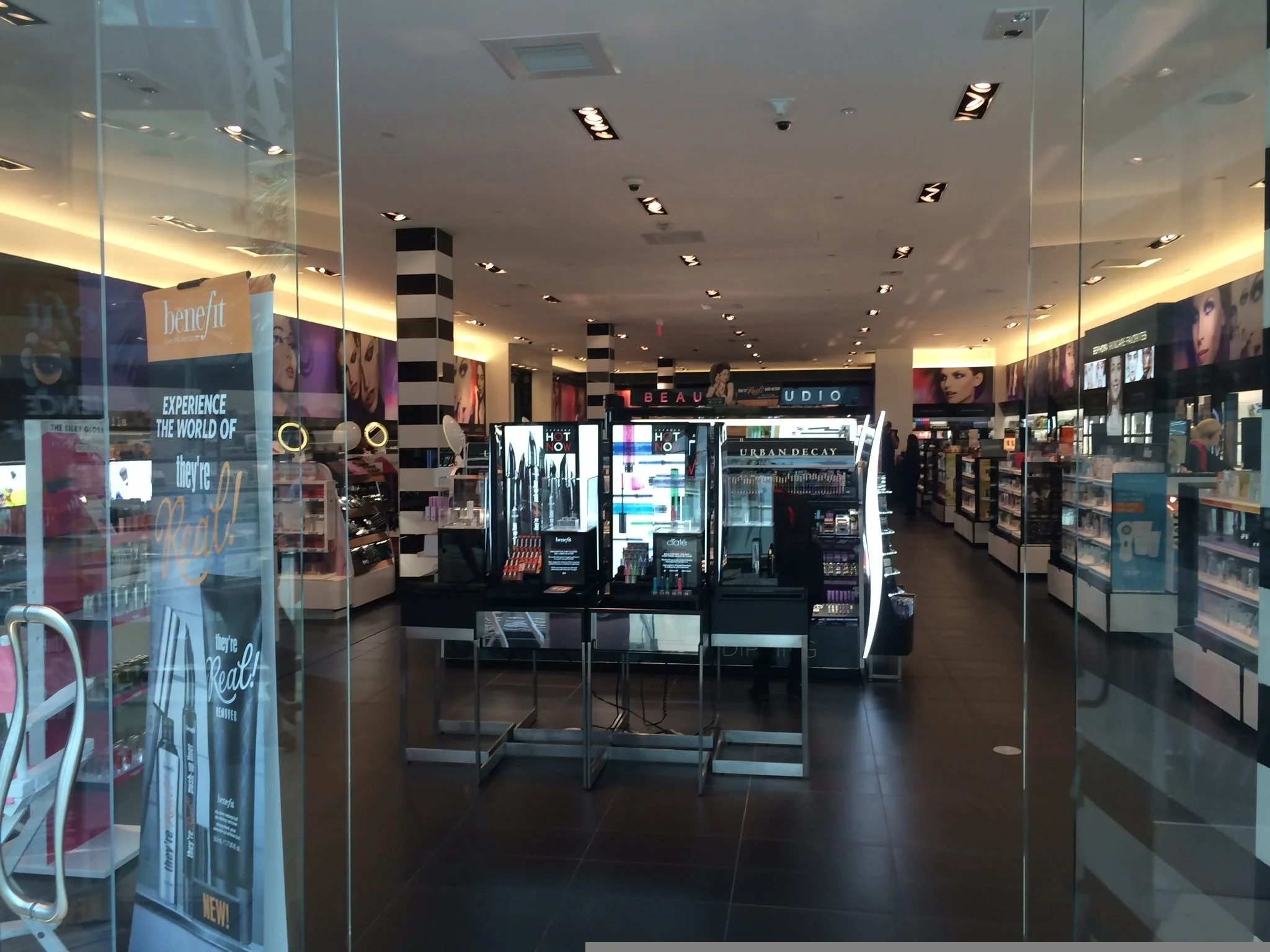 Interior of a beauty store with makeup and skincare products on display, illuminated by ceiling lights, viewed through glass doors.