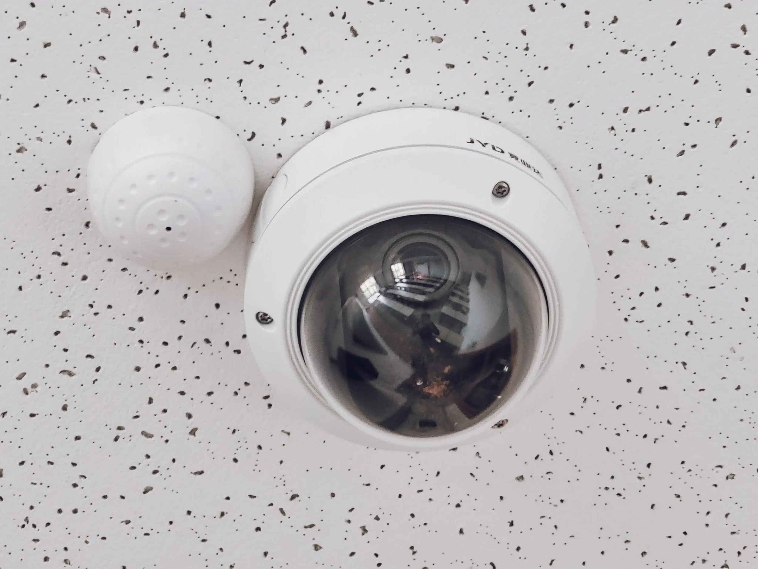 A security camera mounted on a white speckled ceiling with a removable white cover cap nearby.