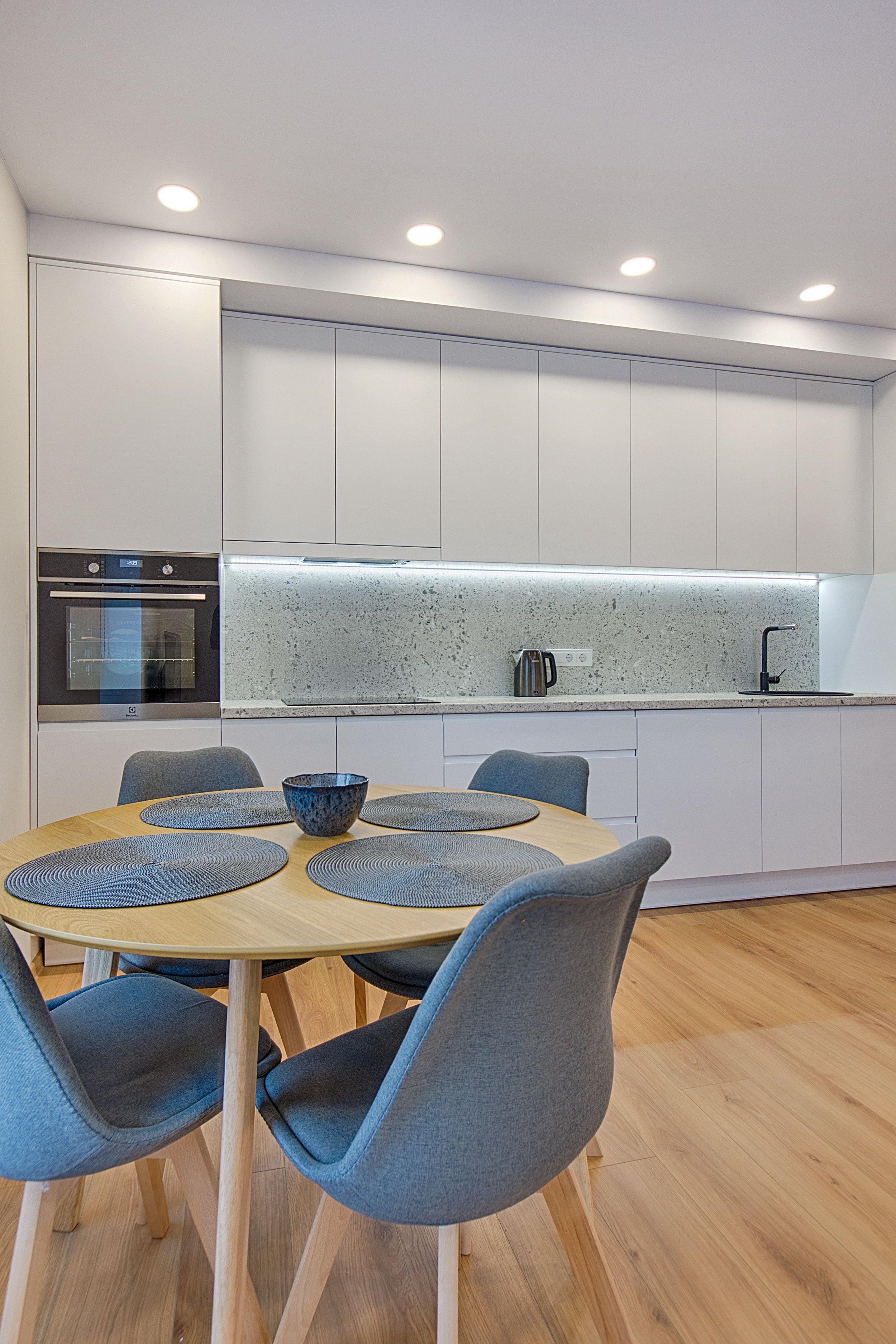 Modern kitchen with white cabinets, speckled backsplash, built-in oven, a countertop with a kettle, and a dining table with chairs and gray placemats.