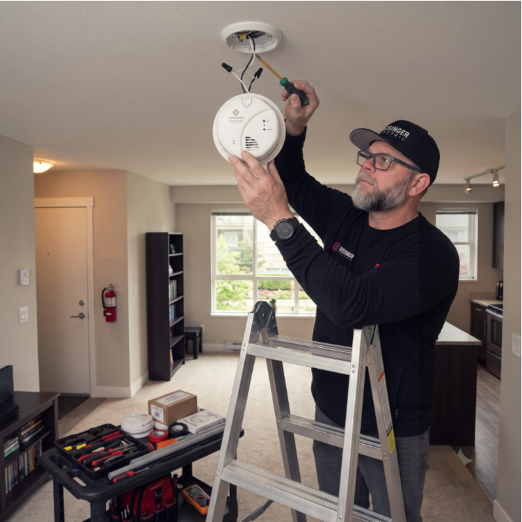 A man installing or maintaining a smoke detector on a ceiling in a living room using a screwdriver while standing on a ladder.