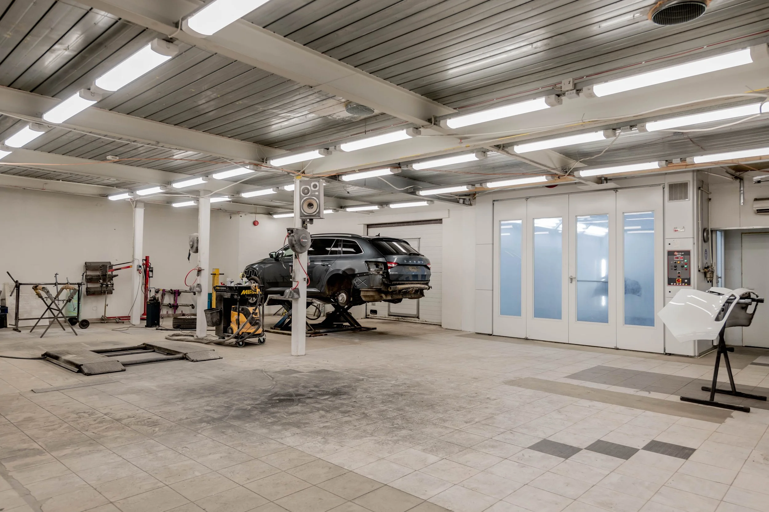 An indoor garage or workshop with a vehicle on a lift, surrounded by various tools and equipment, and bright fluorescent lighting.