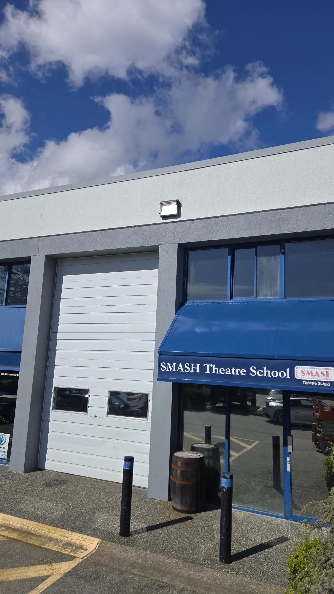 The exterior of SMASH Theatre School building with a blue awning, a white garage door, and a blue sky with clouds.