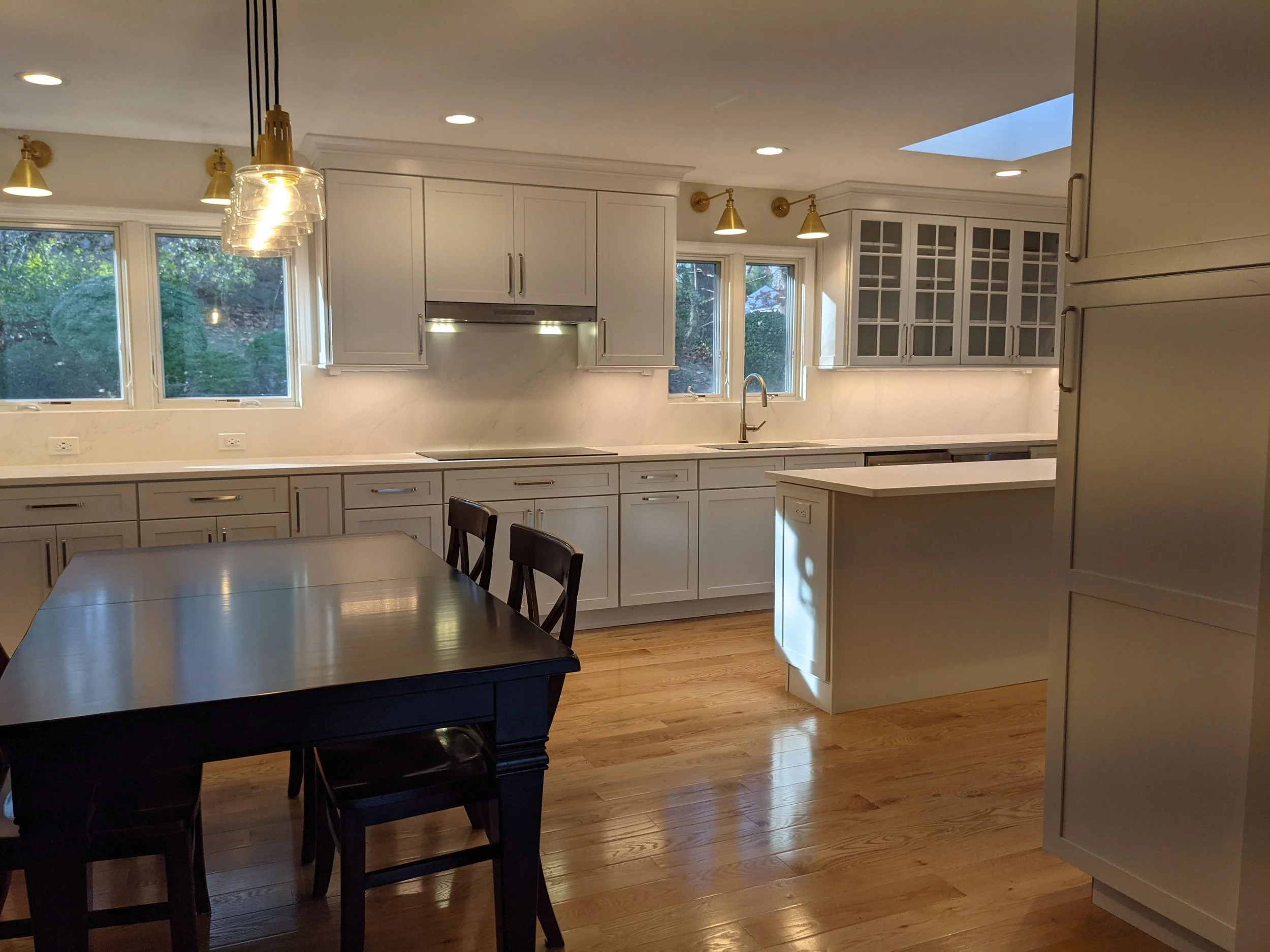 A modern kitchen with white cabinets, a marble backsplash, and large windows. There are pendant lights above the counter, hardwood floors, and a dining table with chairs in the foreground.