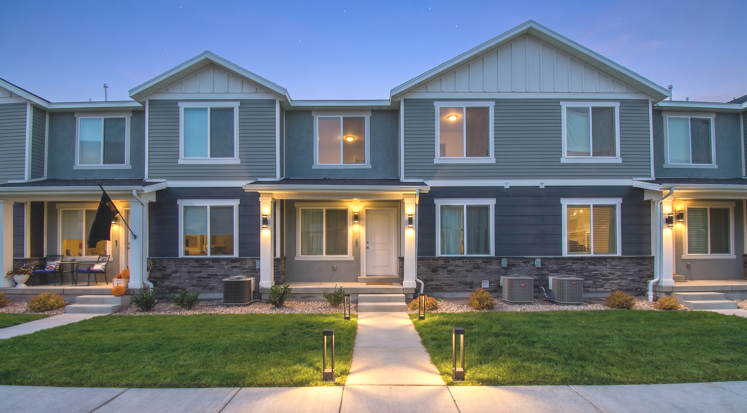 A modern two-story apartment building with gray siding, white trim, and multiple lit windows and exterior lights at dusk. There is a well-maintained lawn with pathway and landscape lighting.