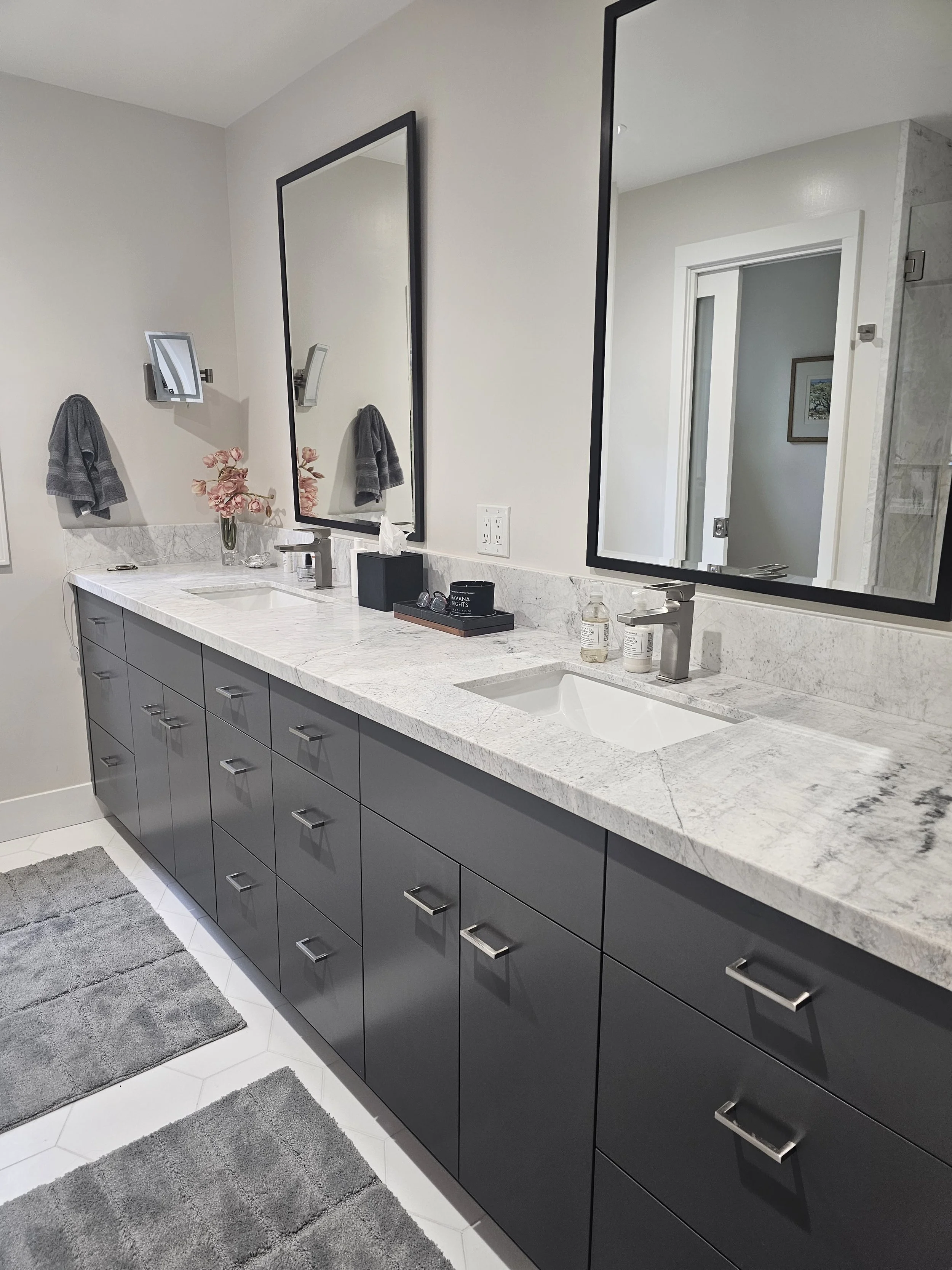 A modern bathroom vanity with a white marble countertop, two sinks, and dark gray cabinets. There are two mirrors above the sinks, each with a black frame. On the countertop, there are bottles of hand soap, a tissue box, and other small items. A gray rug is placed on the white tiled floor in front of the vanity. The walls are painted light, and an open doorway reveals a part of another room with a framed picture.