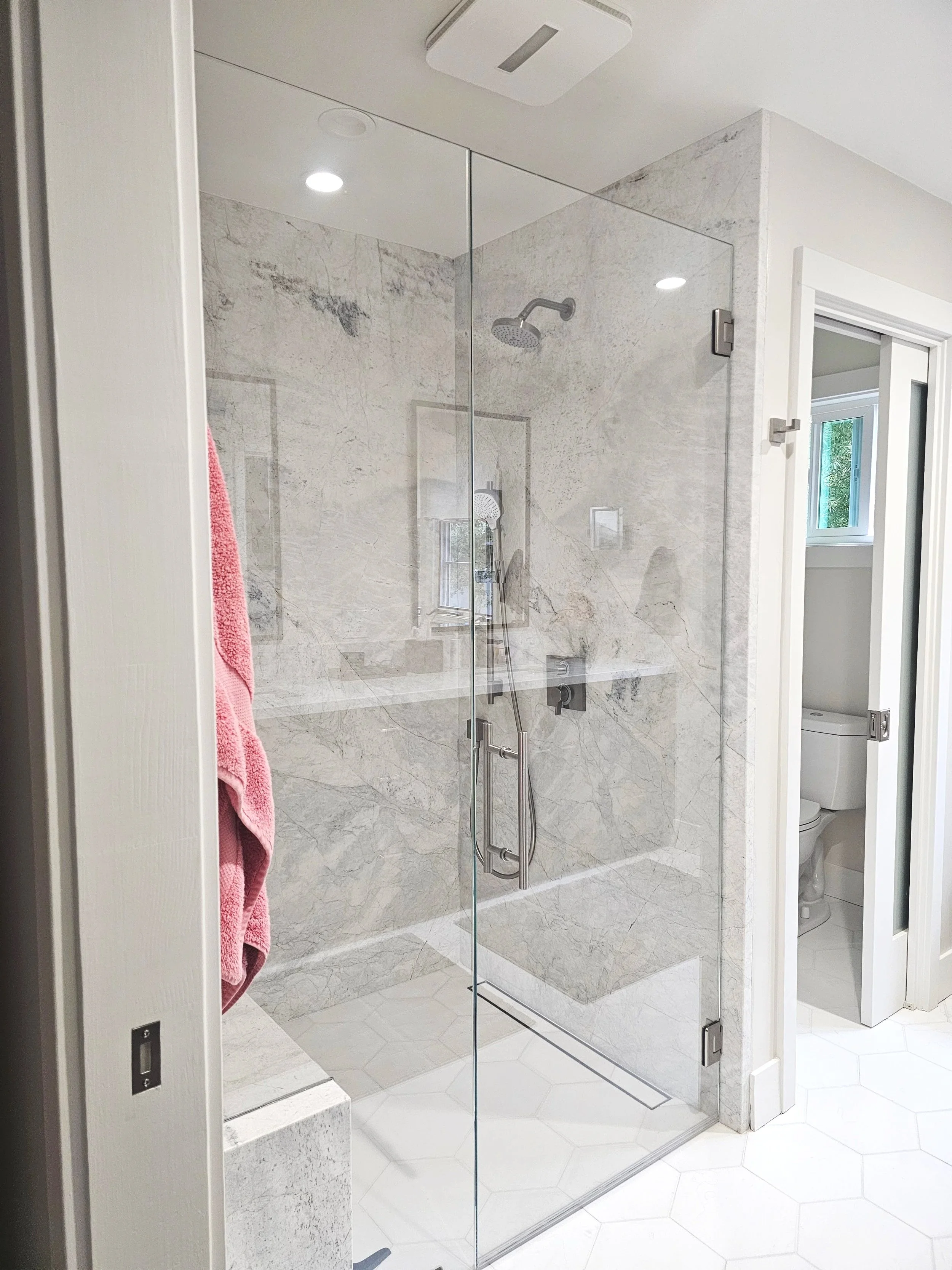 Modern walk-in shower with a glass door, marble tile walls, and a rain shower head, with a pink towel hanging outside and a nearby bathroom toilet visible through a partial door.