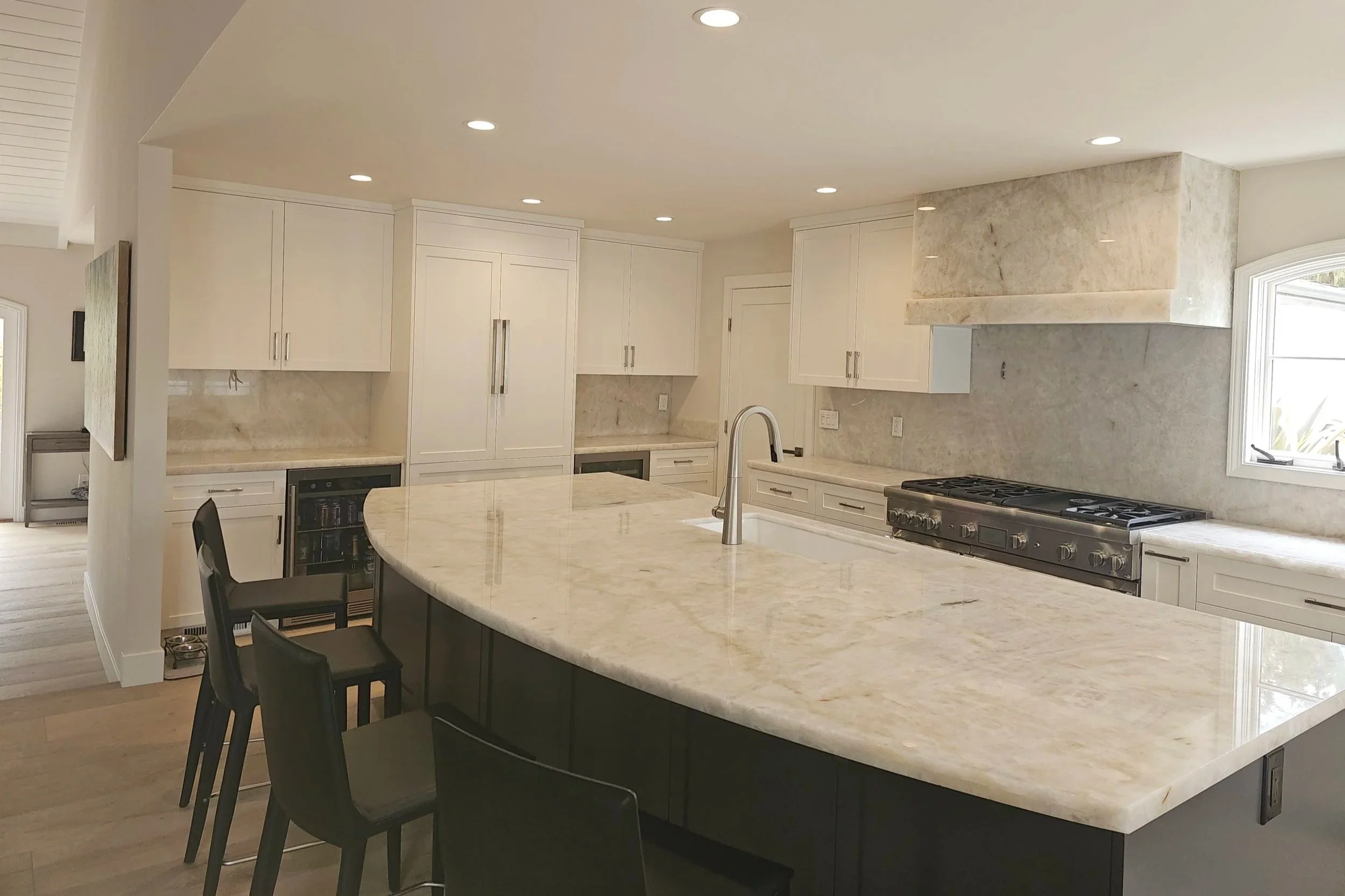 A modern kitchen with white cabinets, marble countertops, and a large kitchen island with seating. There is a stove with an oven on the right and a window on the far right, with recessed lighting in the ceiling.
