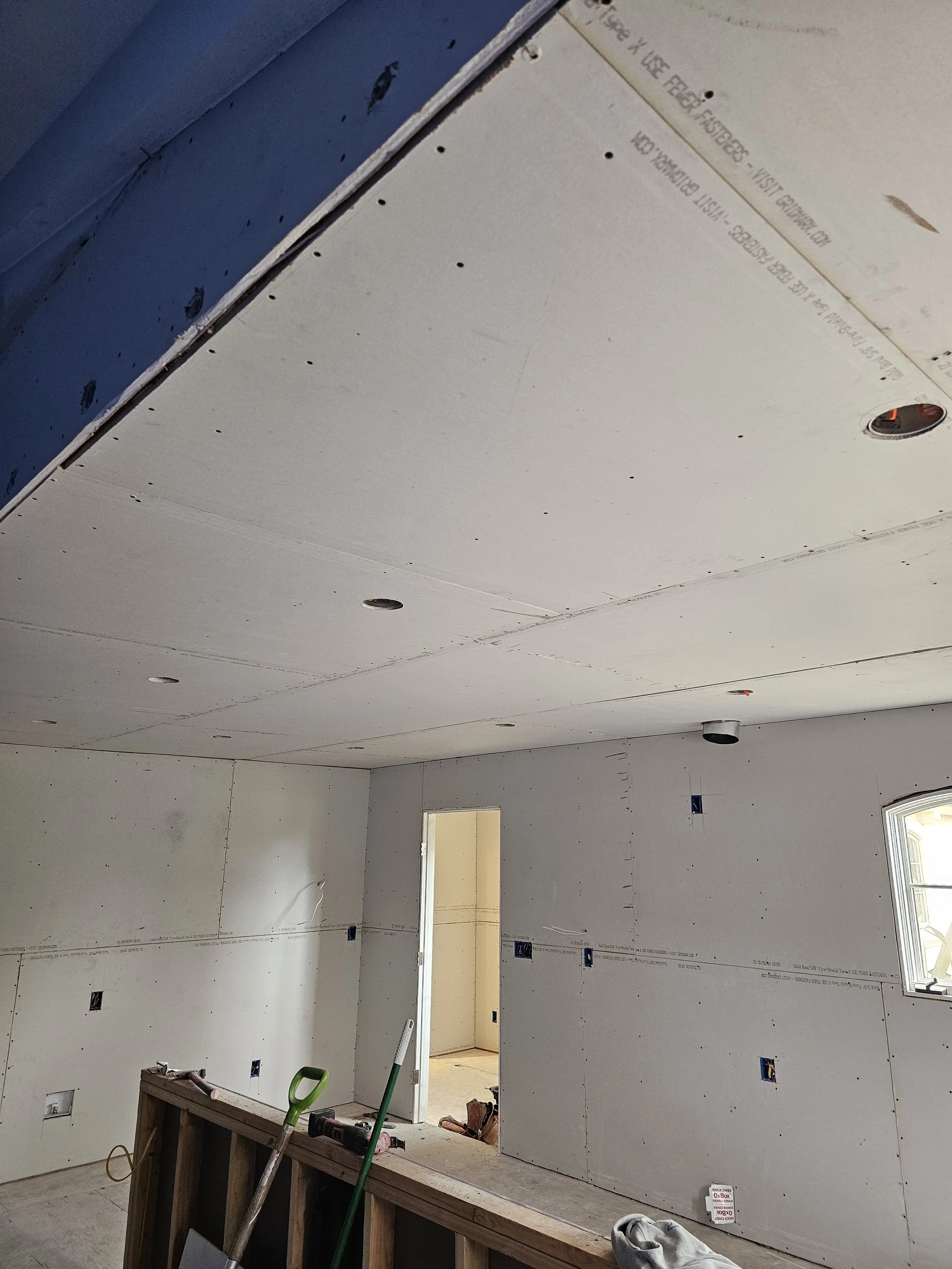 Room under construction with drywall installed on the ceiling and walls, a door opening leading to another space, construction tools and materials like a saw, tools, and green handle in the foreground.