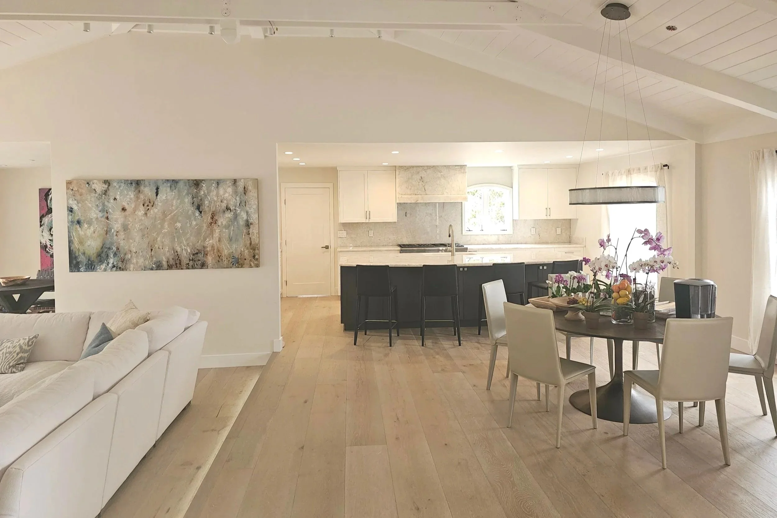Open-concept living area with white sofa, dining table with chairs, and kitchen in the background, featuring white cabinets and a marble backsplash, illuminated by natural light from windows.
