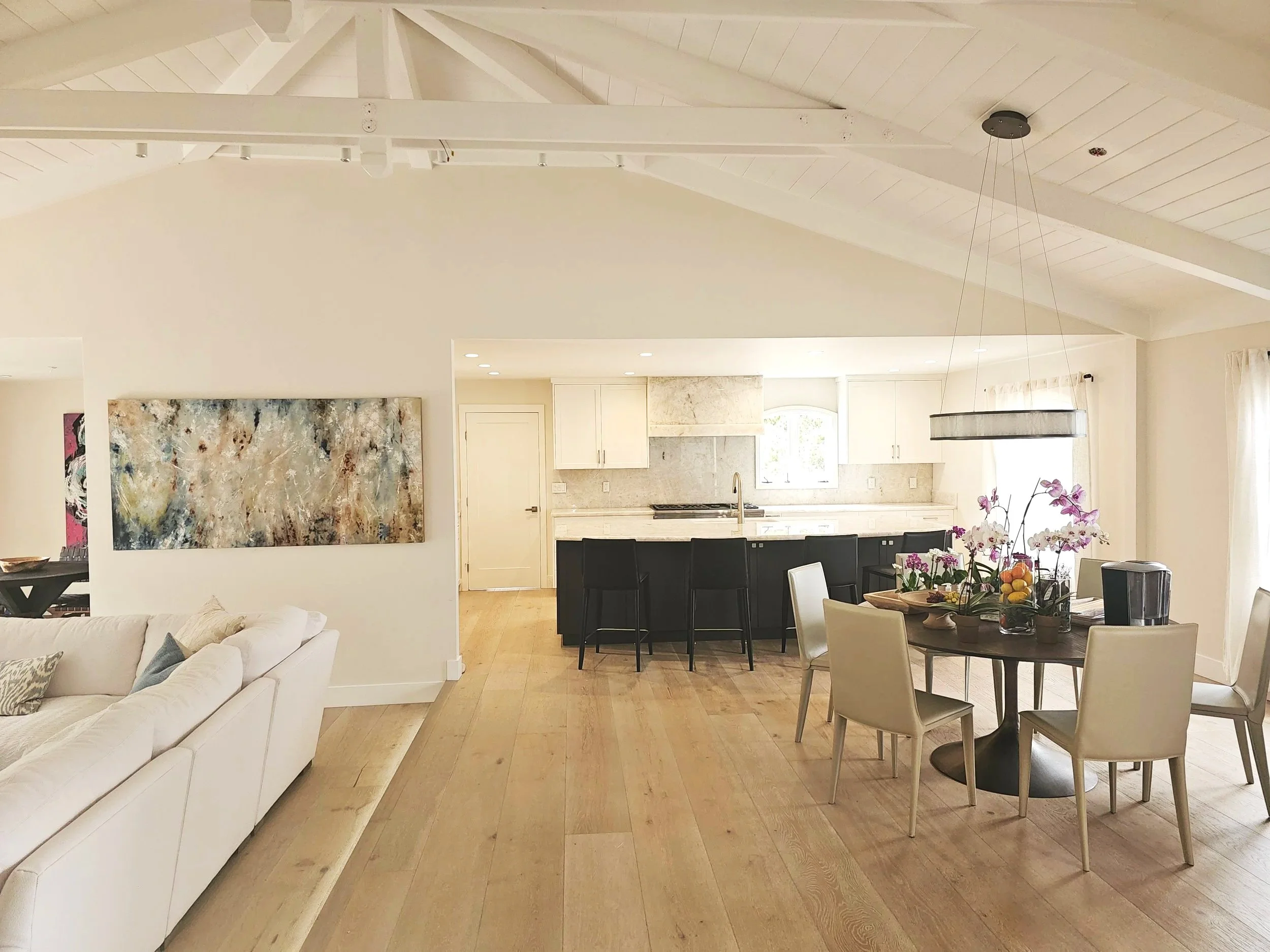 Interior view of a modern, open floor plan kitchen and dining area with white walls, wooden flooring, and high, vaulted ceiling with white beams. The kitchen has white cabinets, marble backsplash, and a large window. The dining table is round with beige chairs, topped with a fruit bowl and orchid flowers. A white sofa with decorative pillows is visible in the foreground.