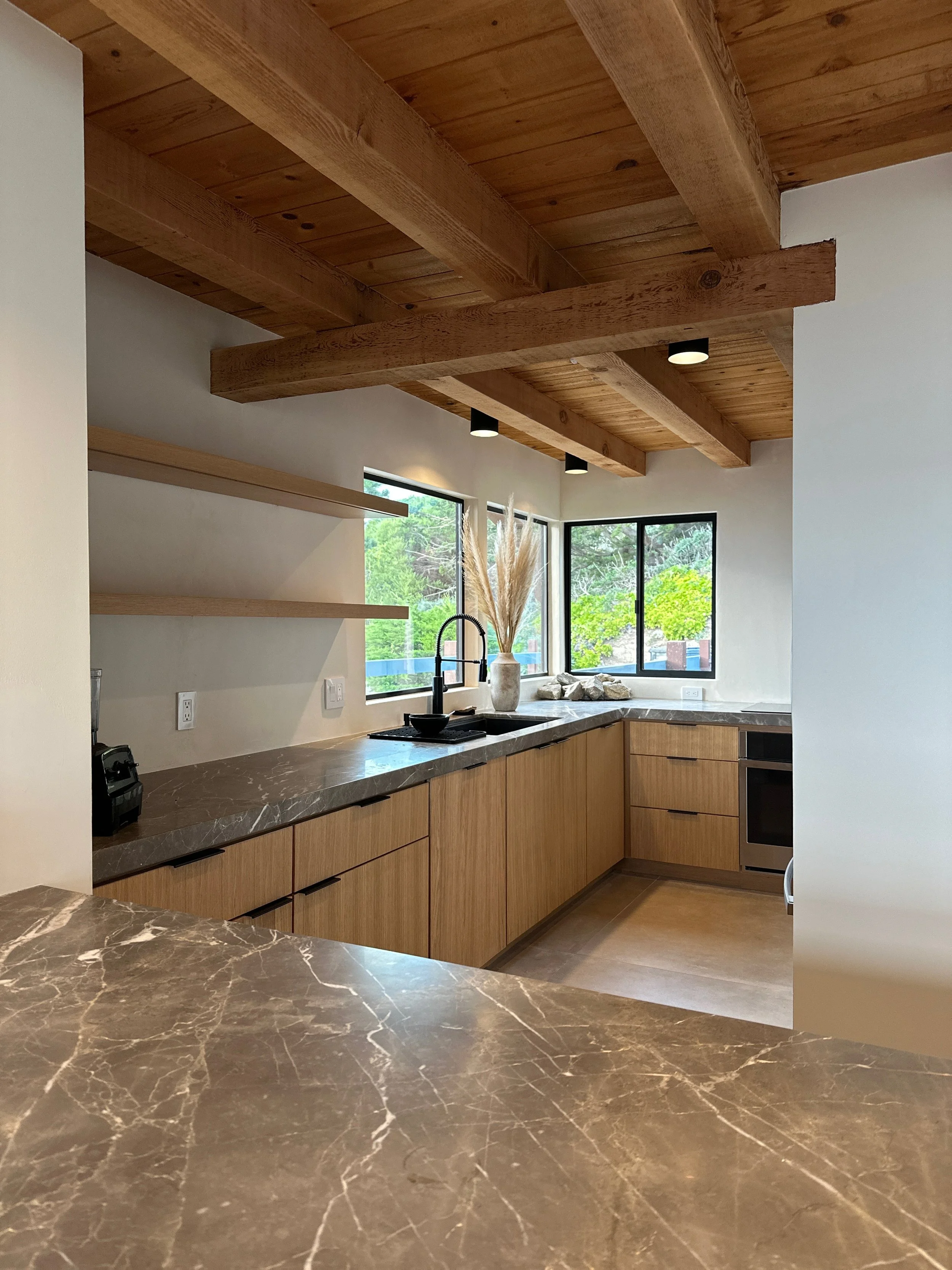 Modern kitchen with wooden ceiling beams and a marble countertop, large windows with a view of green trees, and a vase with dried plants on the counter.