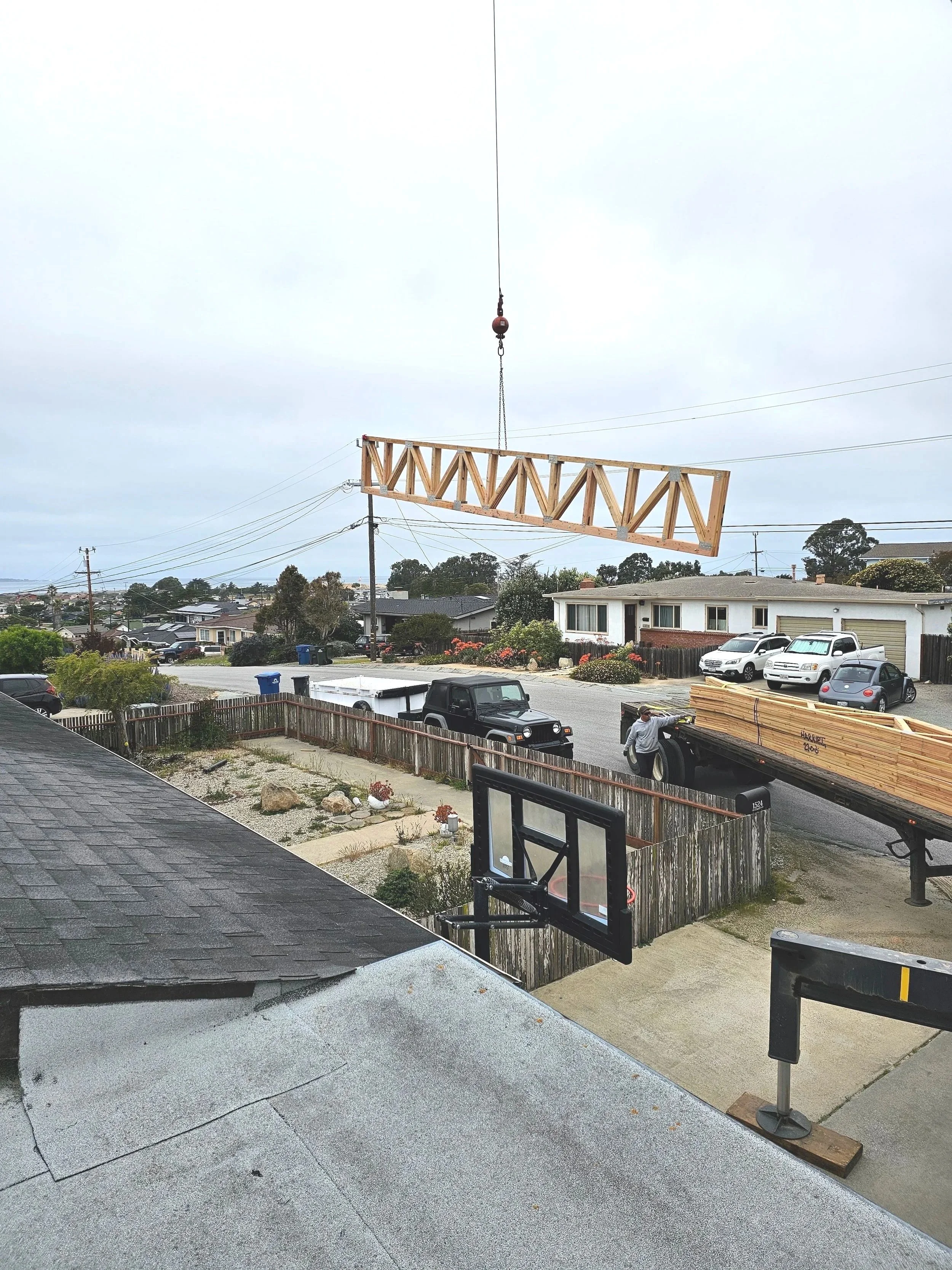 A residential neighborhood with a wooden porch railing, a basketball hoop, a truck carrying lumber, and houses in the background. A crane is lifting a wooden structure.
